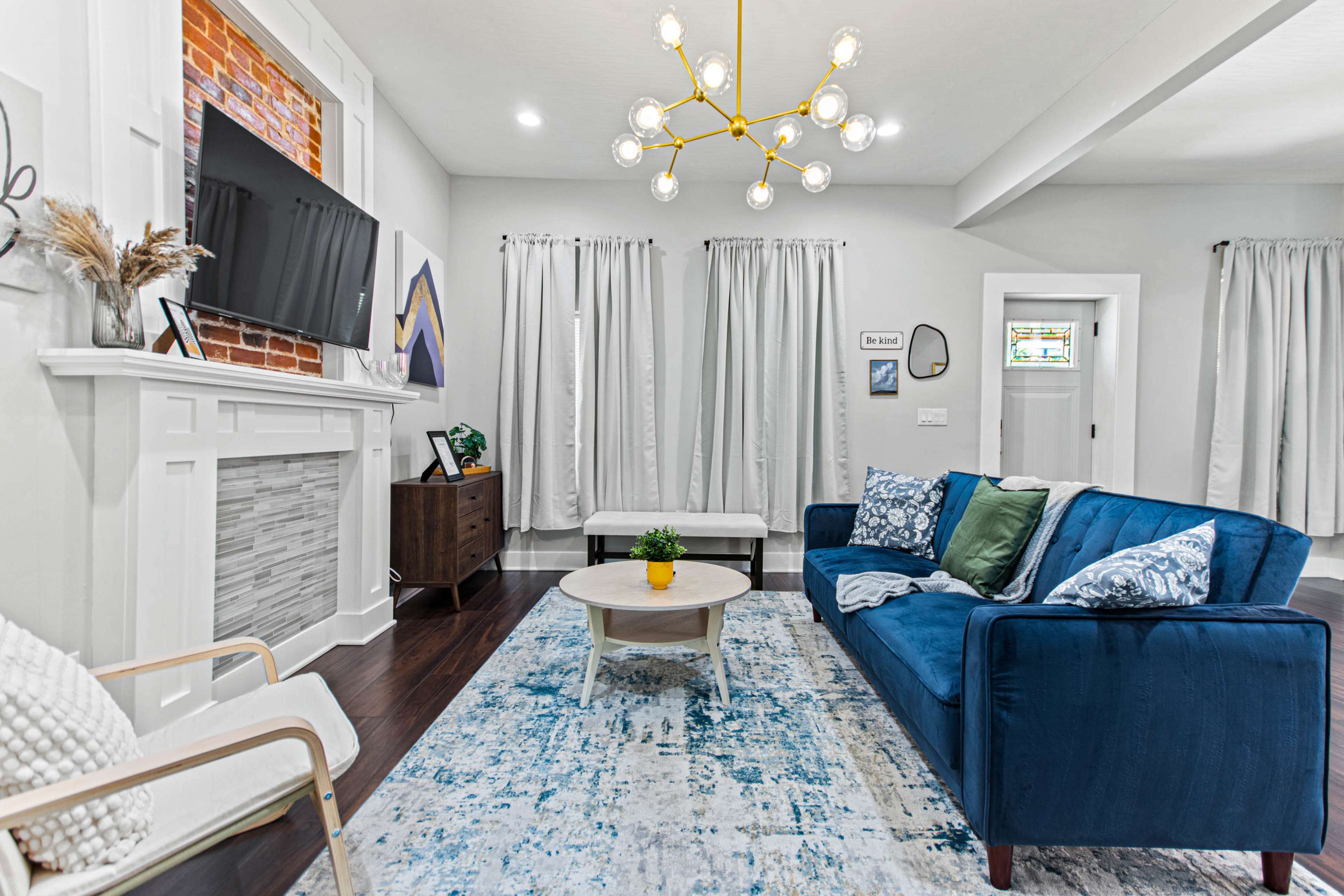 A modern living room features a blue couch, a round coffee table, a mounted television, and decorative curtains framing a window.