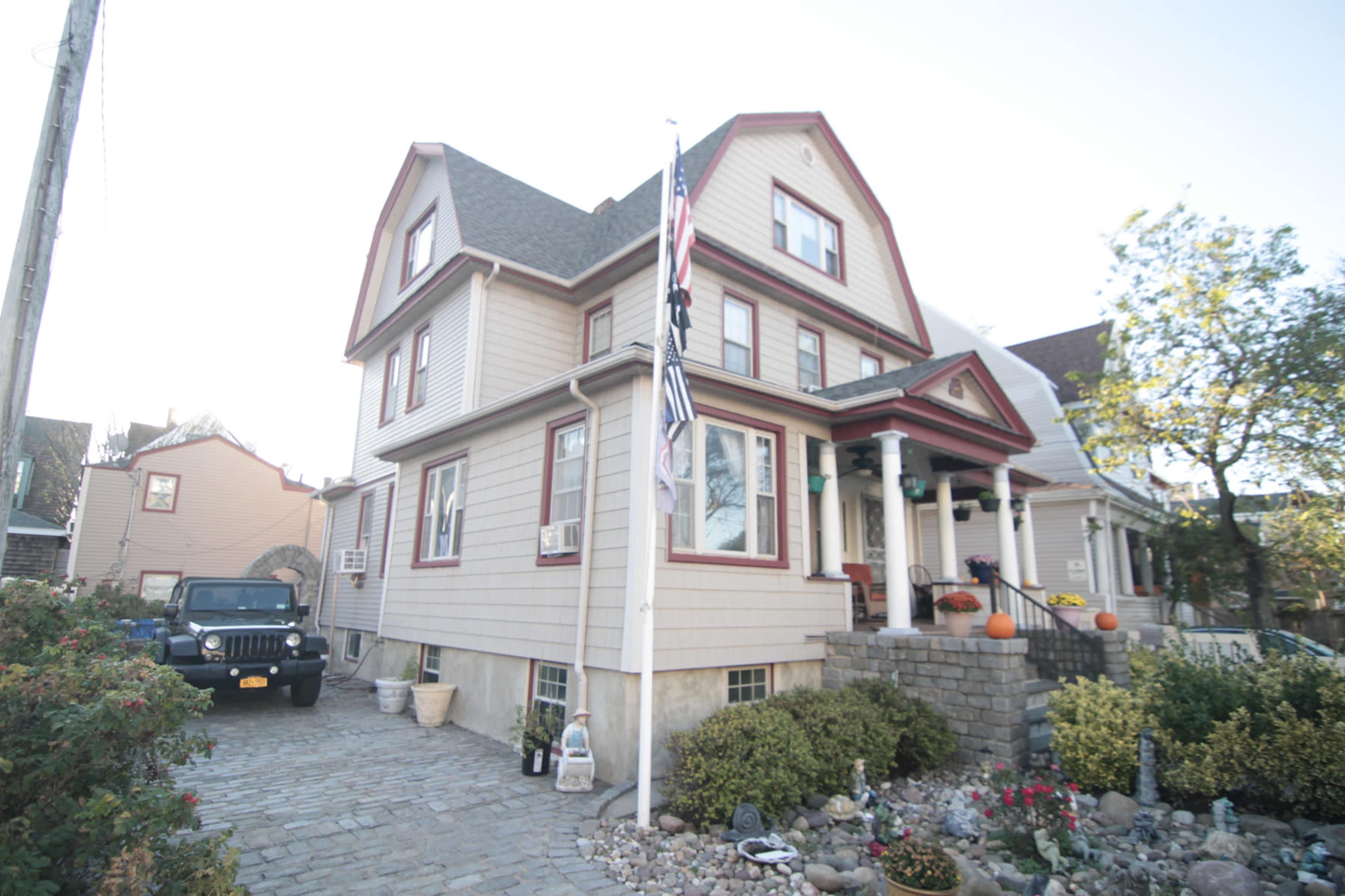 A large, two-story house with a gabled roof and a stone pathway is situated next to a parked black Jeep, featuring a flagpole with an American flag in front.