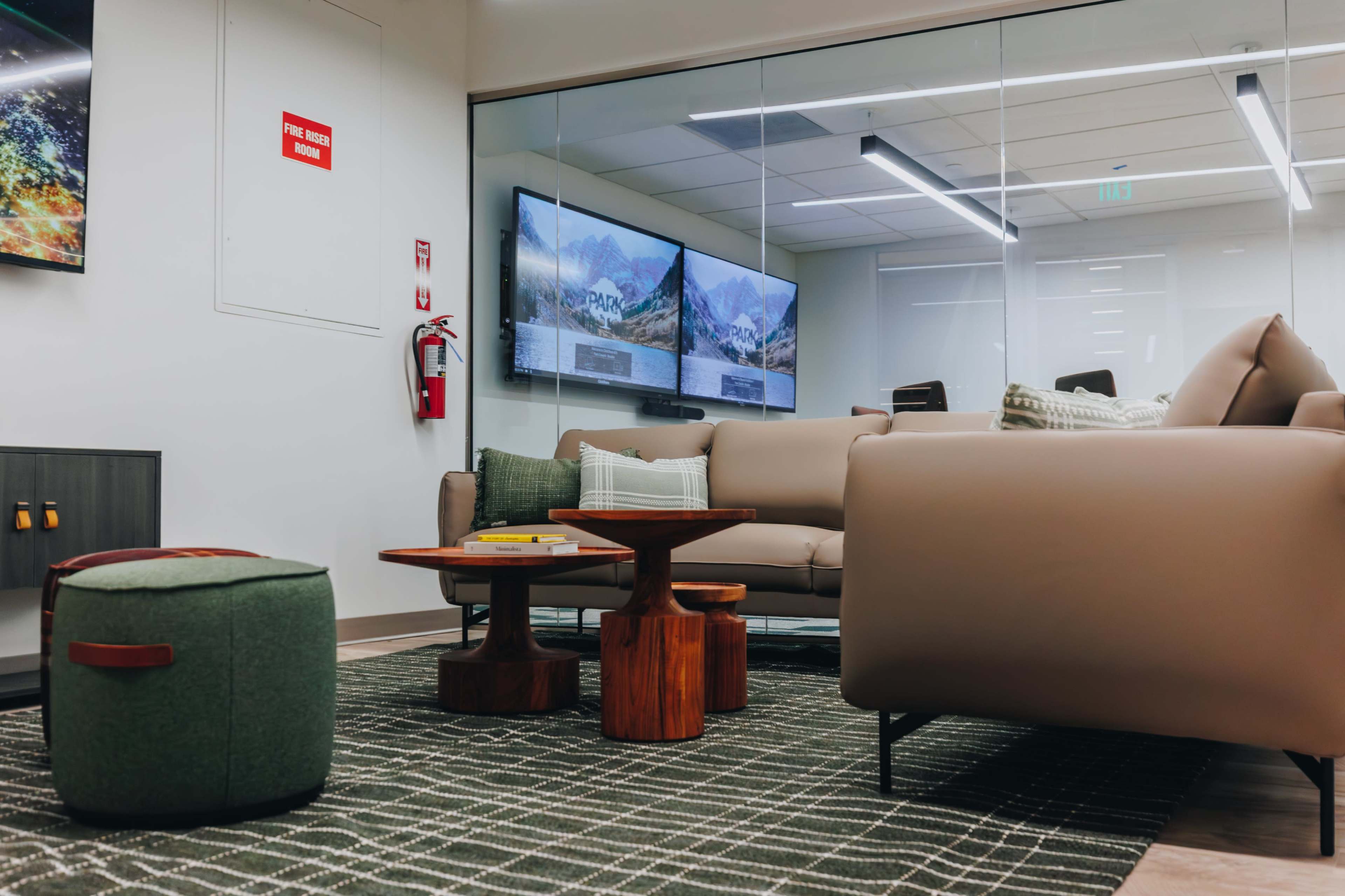 The image shows a modern lounge area with a beige couch, wooden tables, a green pouf, and large screens displaying mountains against a glass wall.
