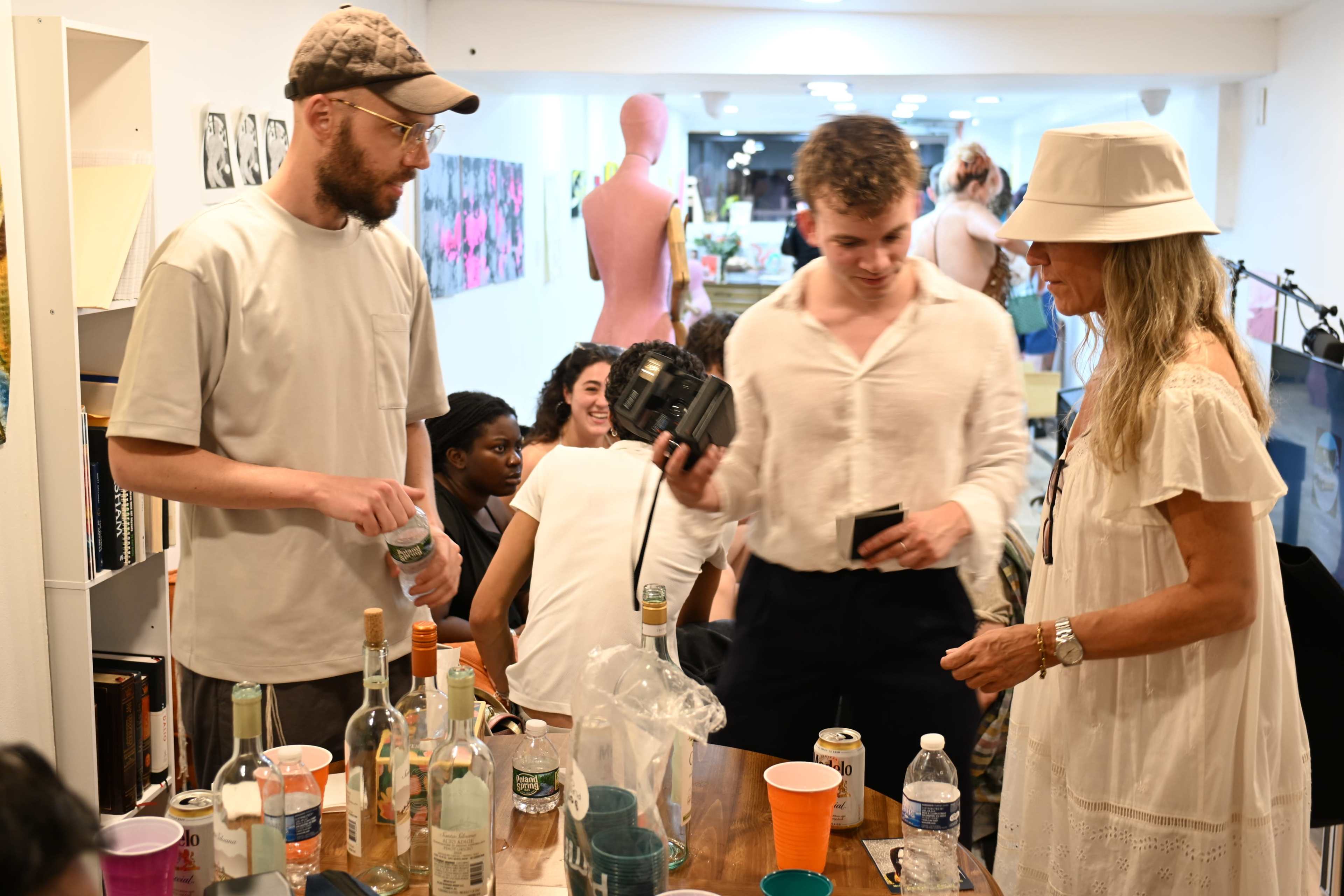 A group of people interacts around a table cluttered with empty bottles and drinks in a lively indoor setting.