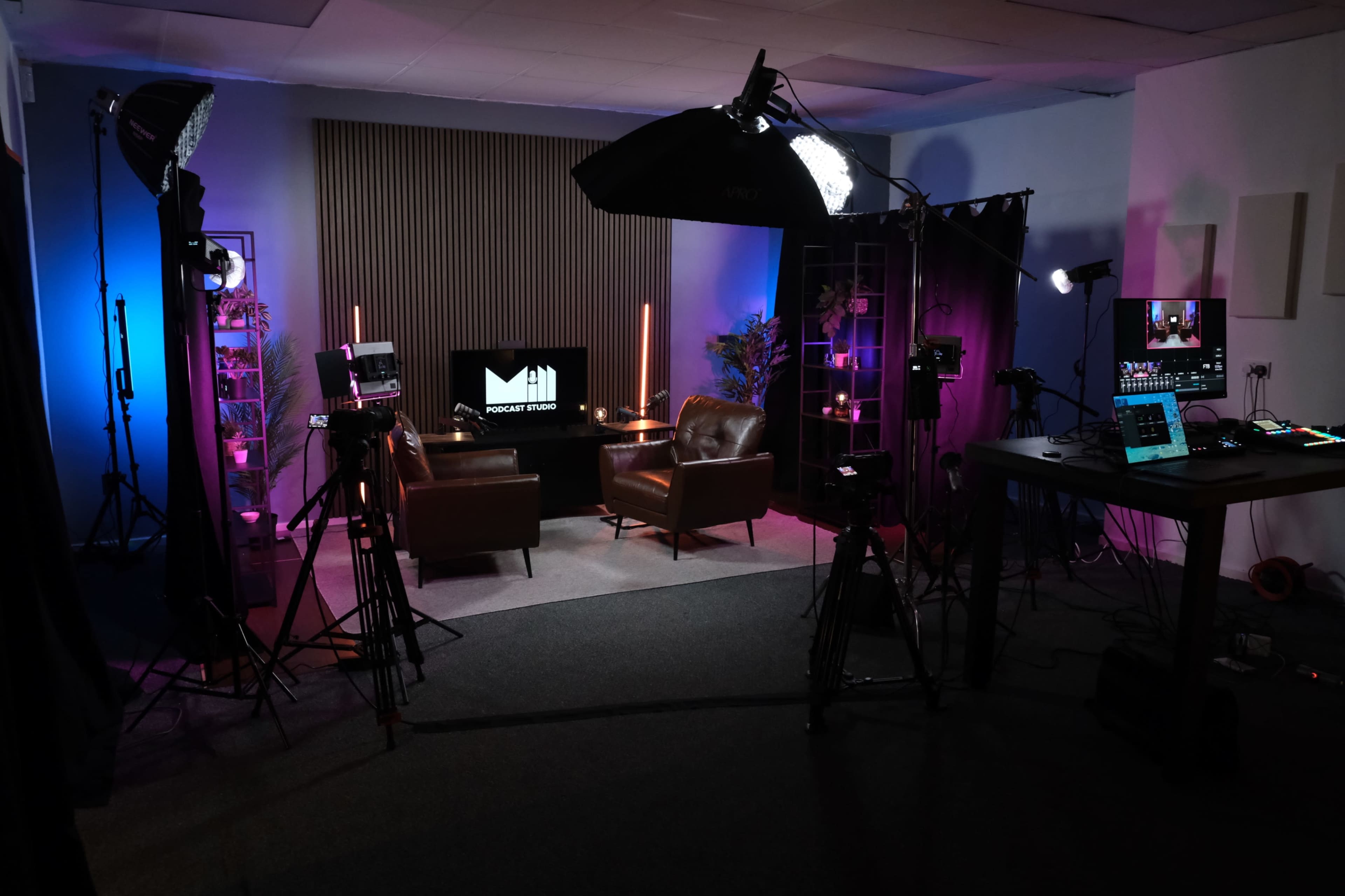 The image shows a podcast studio setup with two leather chairs, a television screen displaying a logo, various camera equipment, and colorful lighting.