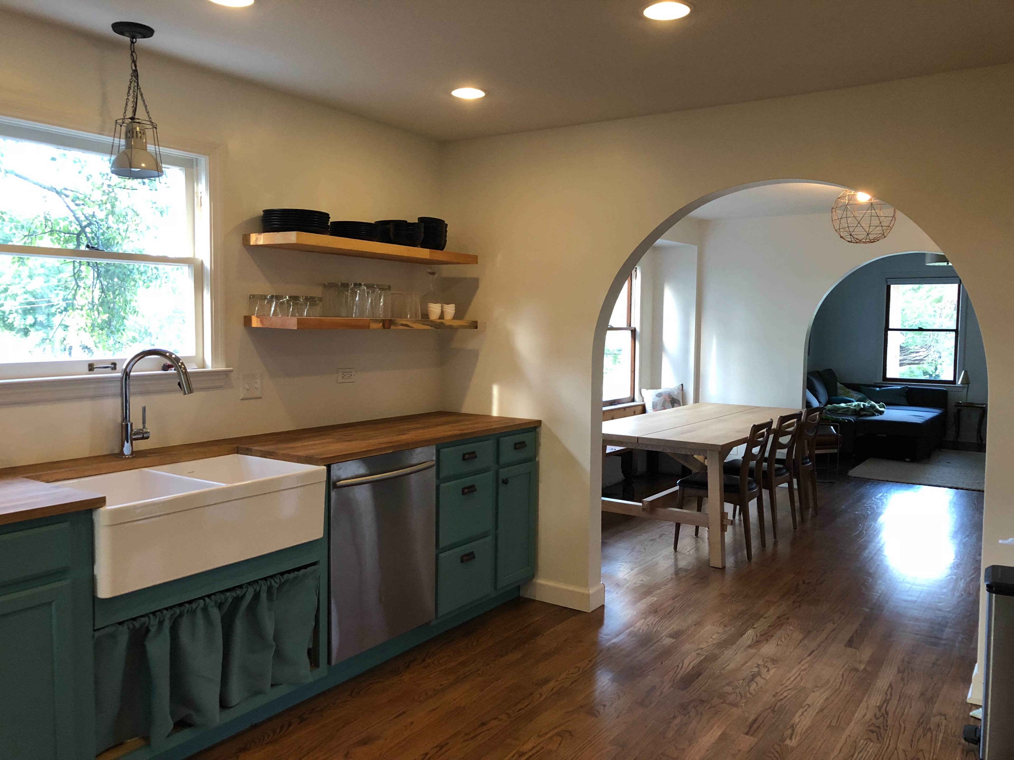 A kitchen with a farmhouse sink and wooden countertops opens to a dining area with a table and chairs, framed by a large archway.