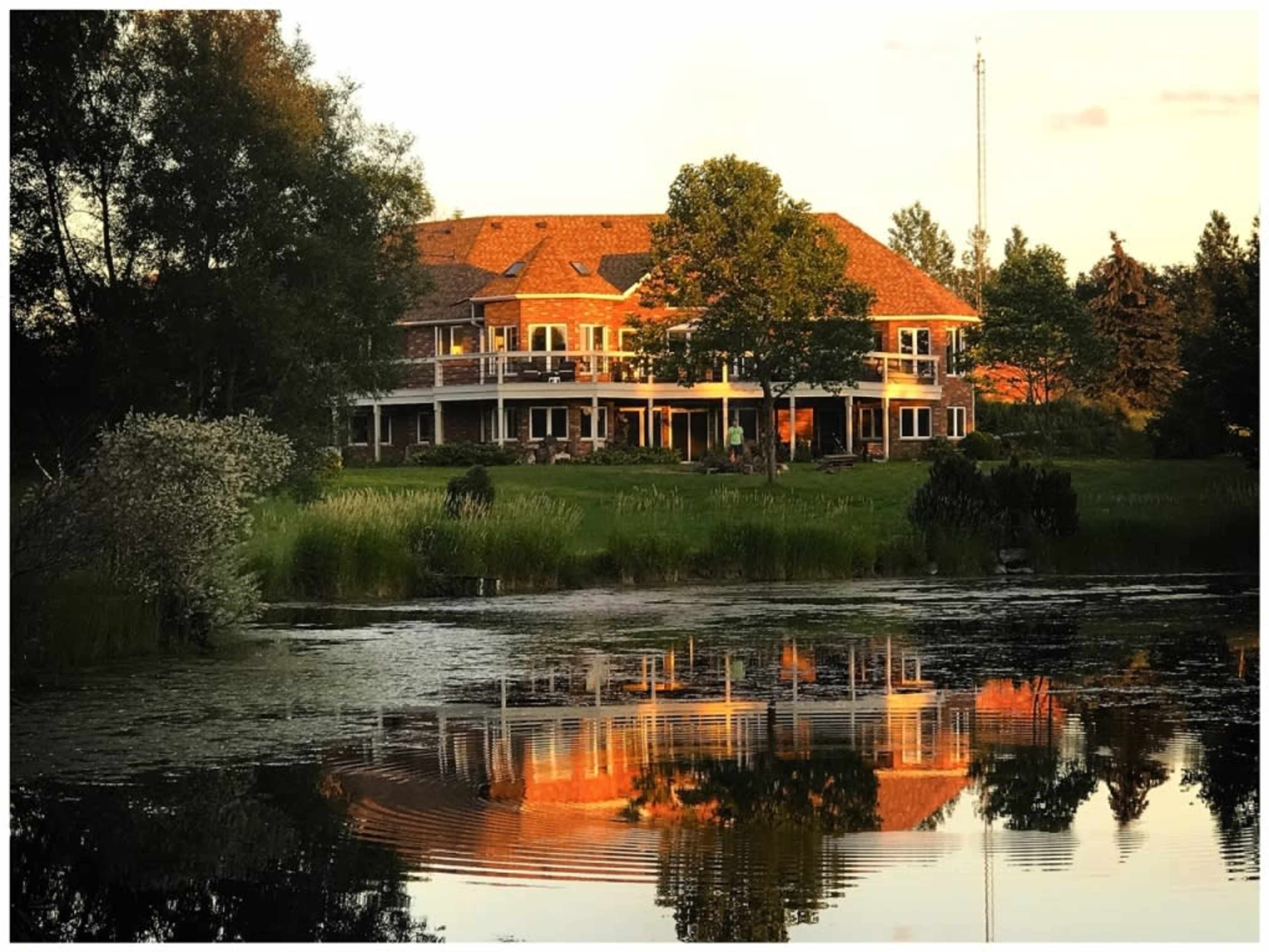 A large, two-story brick house is reflected in a calm pond surrounded by greenery.