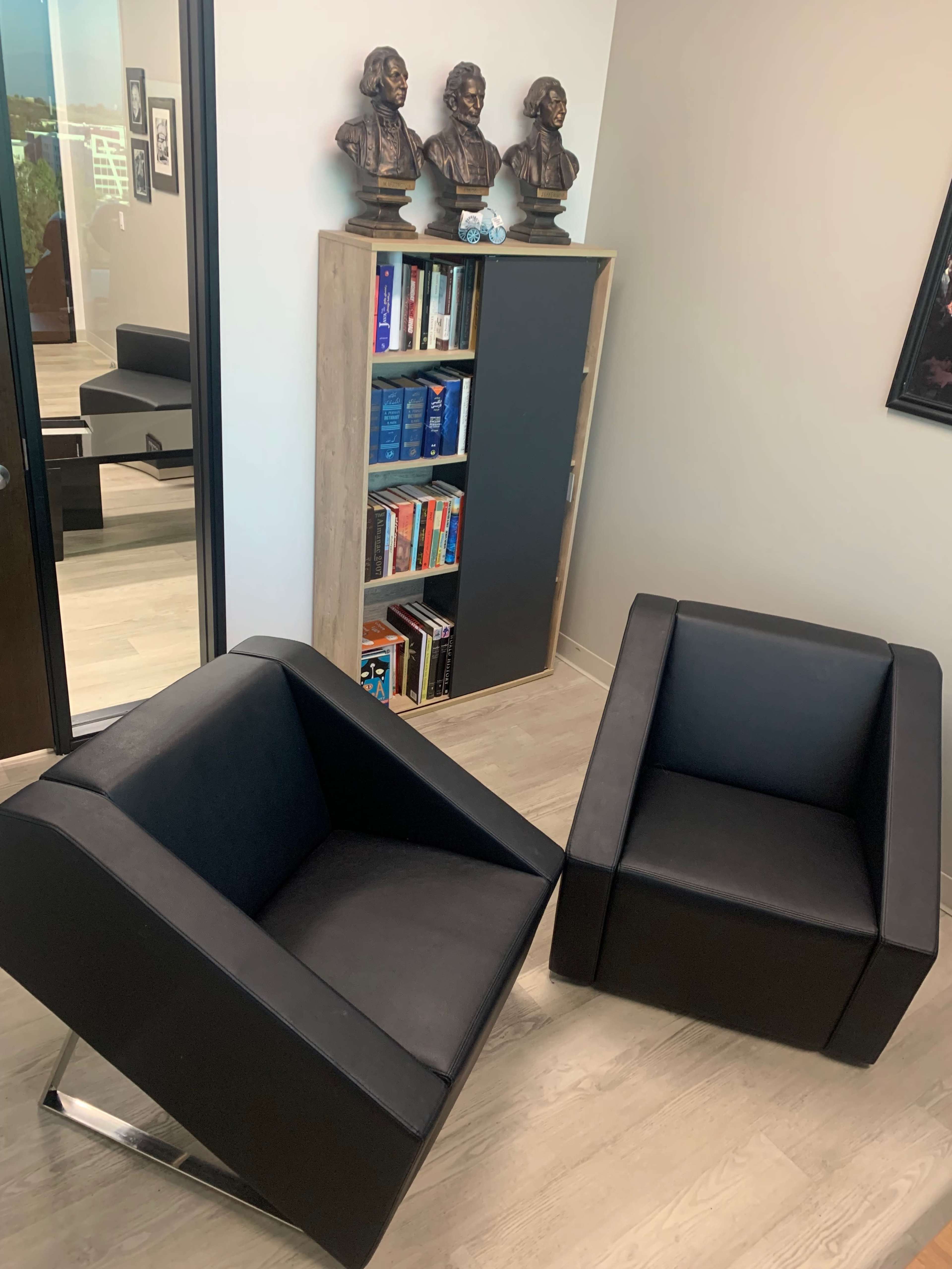 A modern office space featuring two black chairs and a bookshelf filled with books, along with a bust display of three figures.