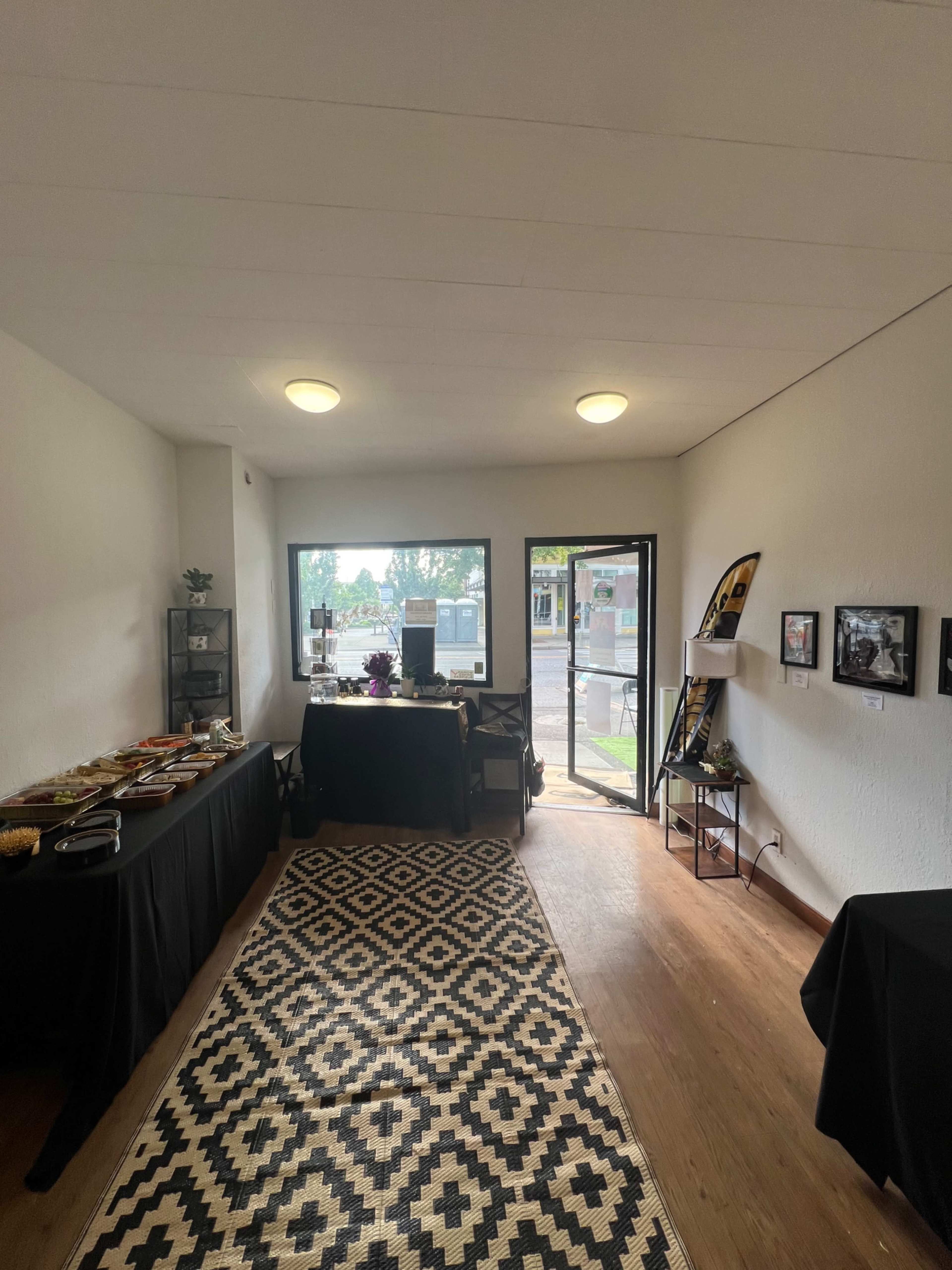 The image shows a neatly arranged interior space with black tables displaying items, a large window, and an entrance door leading outside.