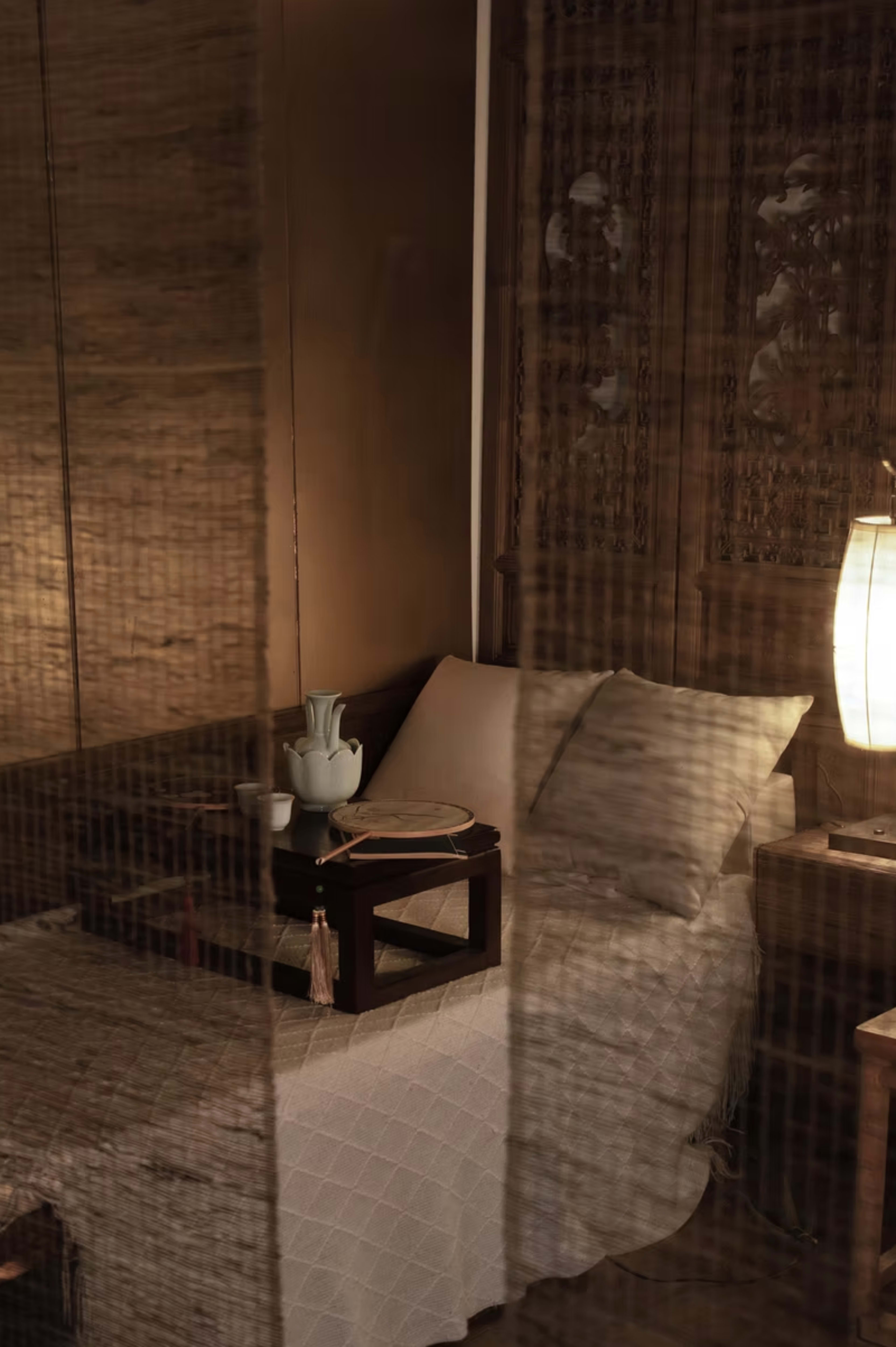 A minimalistic bed with a wooden headboard is set against a softly lit wall, accompanied by a small bedside table and a lamp.
