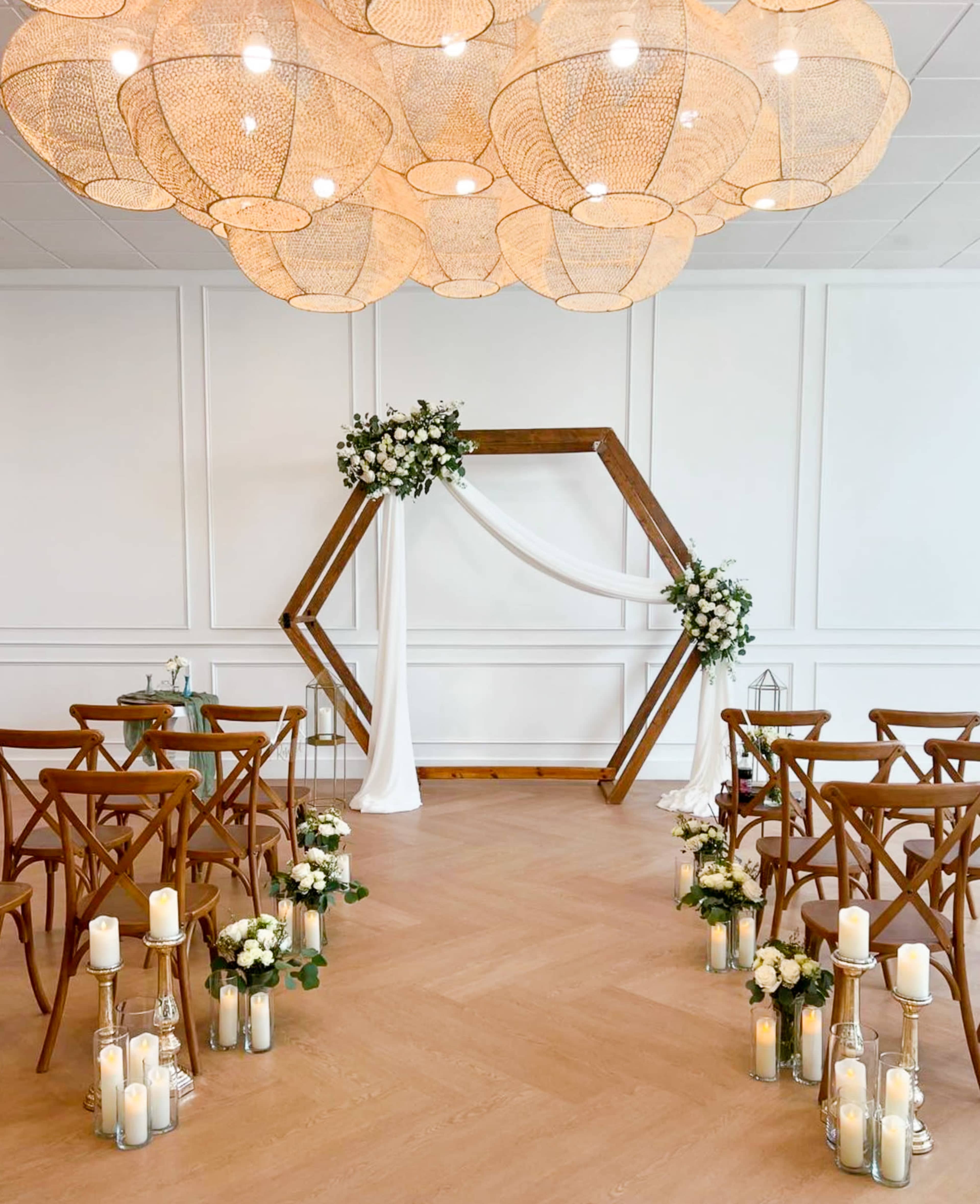 A wedding ceremony setup features an arched wooden structure adorned with white drapery and flowers, flanked by rows of wooden chairs and candles along the aisle.