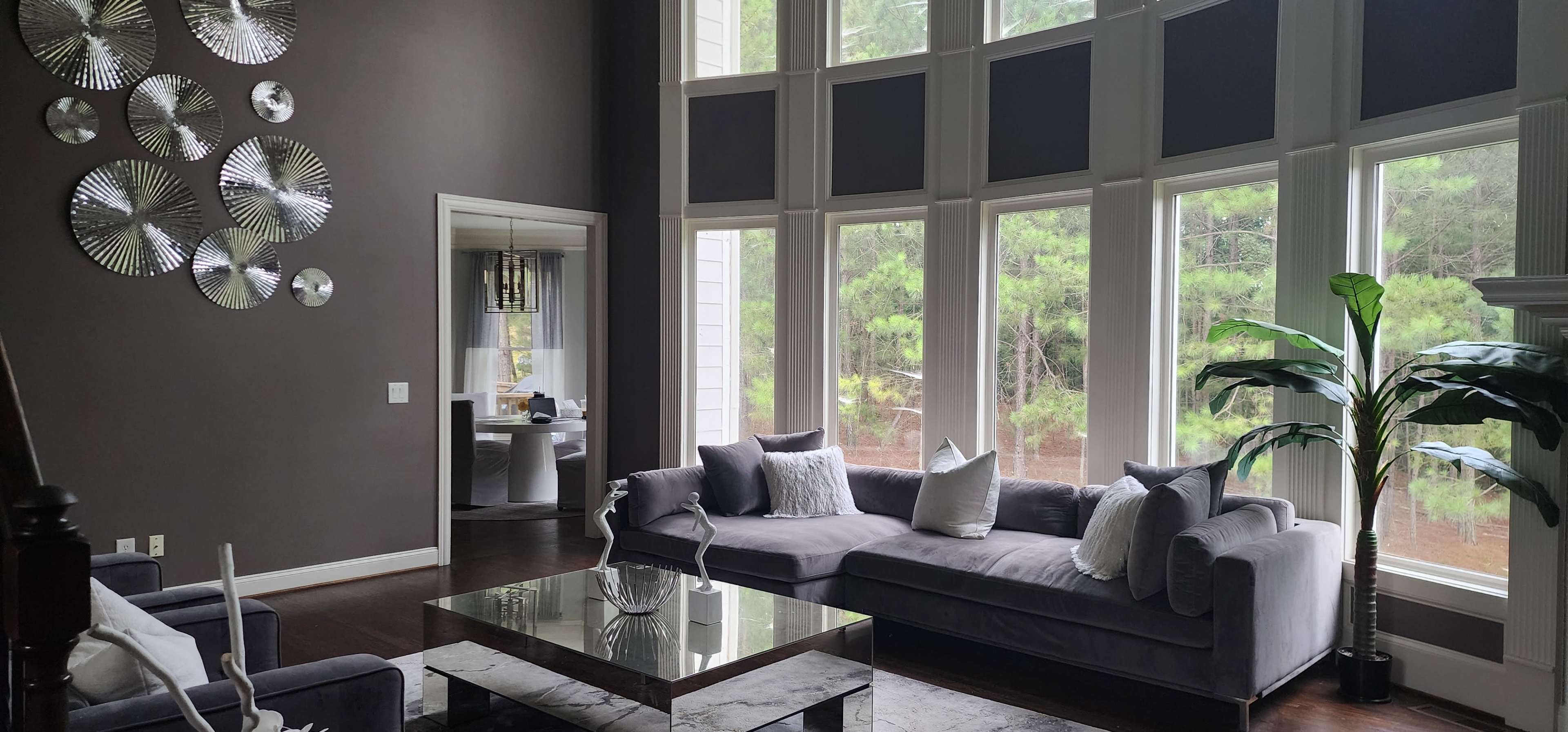 The image shows a modern living room with large windows, a gray sectional sofa, a glass coffee table, and decorative wall art made of circular mirrors.
