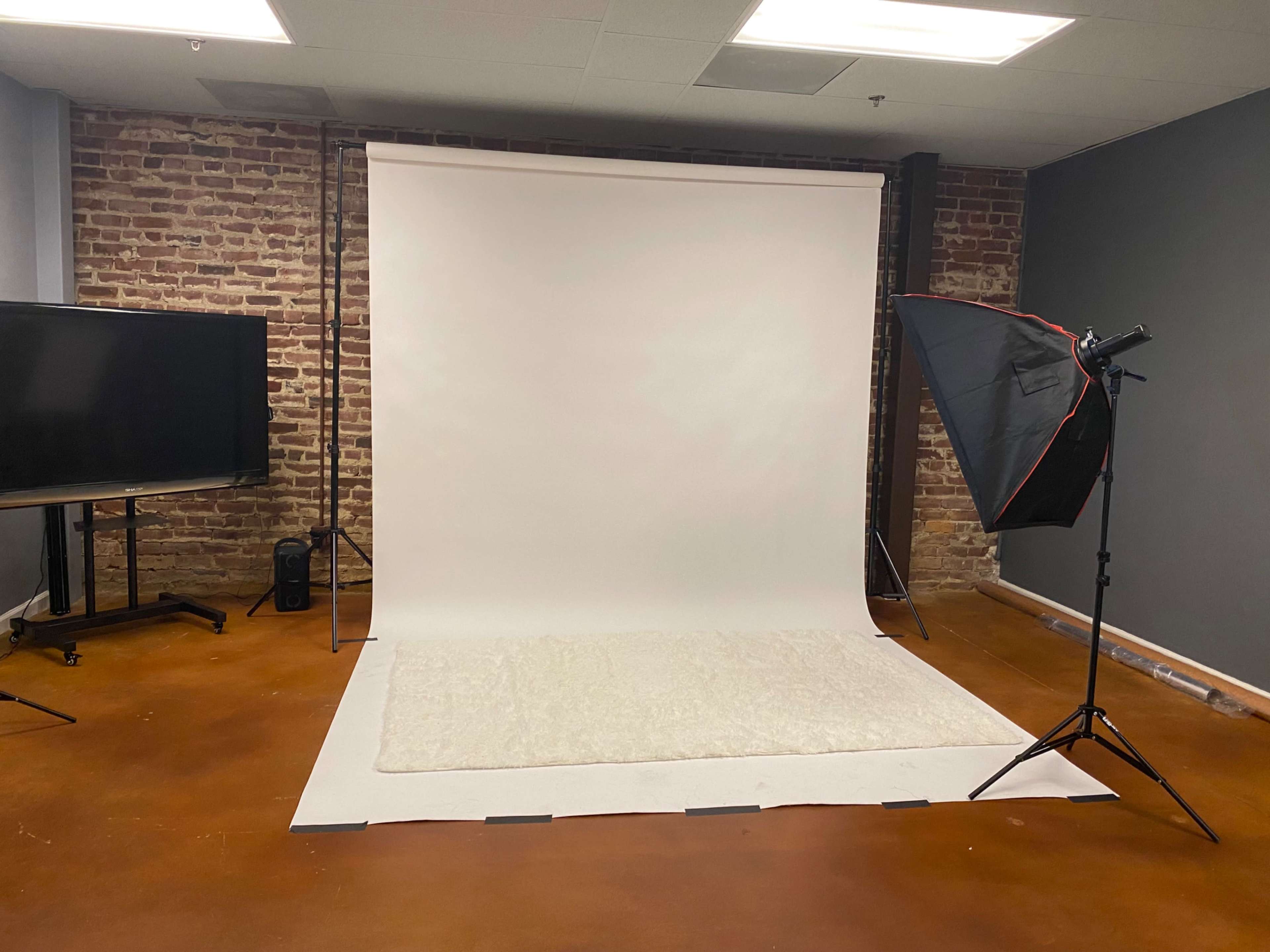 The image shows a photography studio with a white backdrop, a soft rug on the floor, a light stand, and a television screen against a brick wall.
