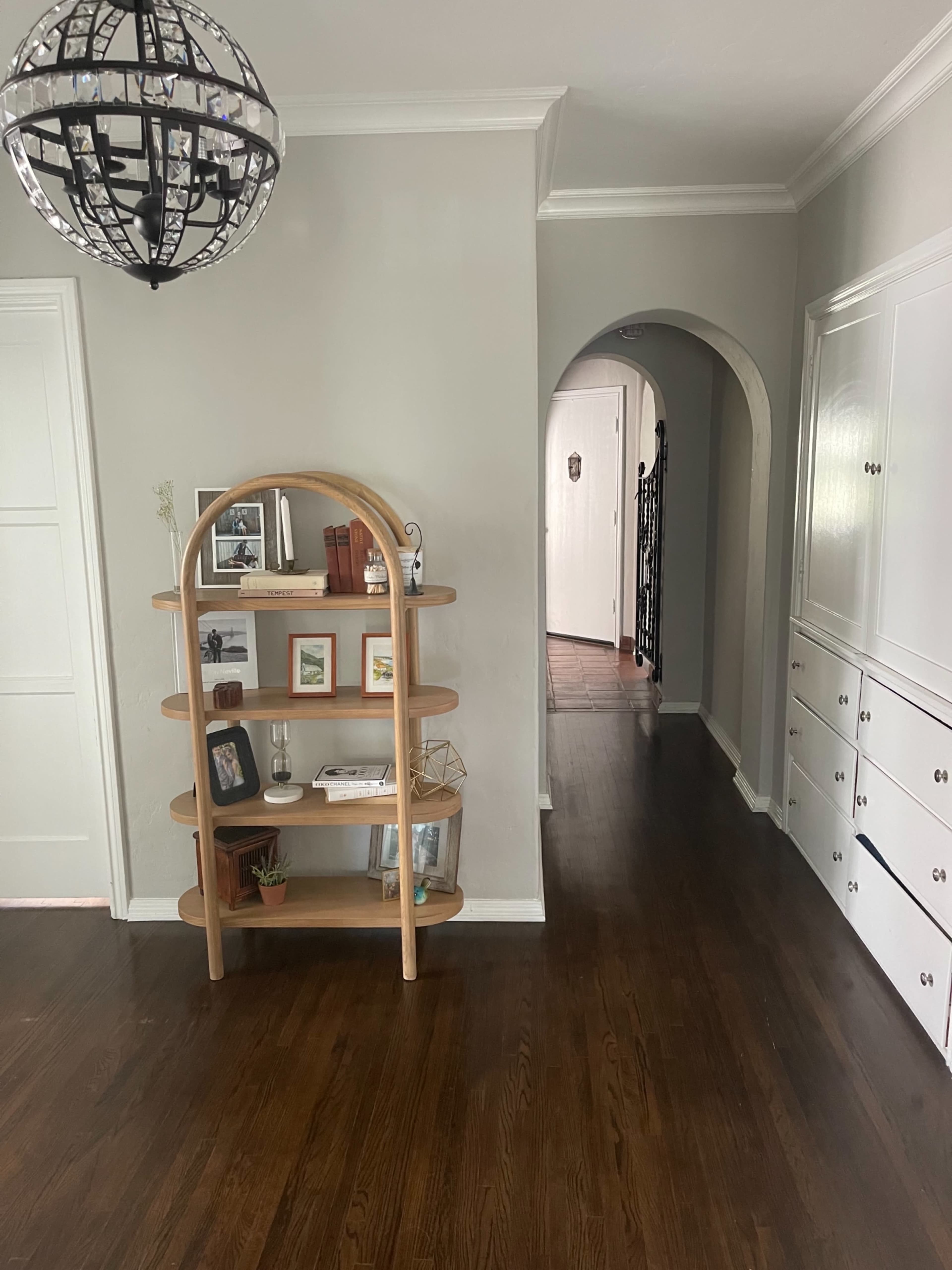 The image shows a well-lit hallway with a wooden shelf holding books and decorative items, leading to an arched doorway and a staircase.