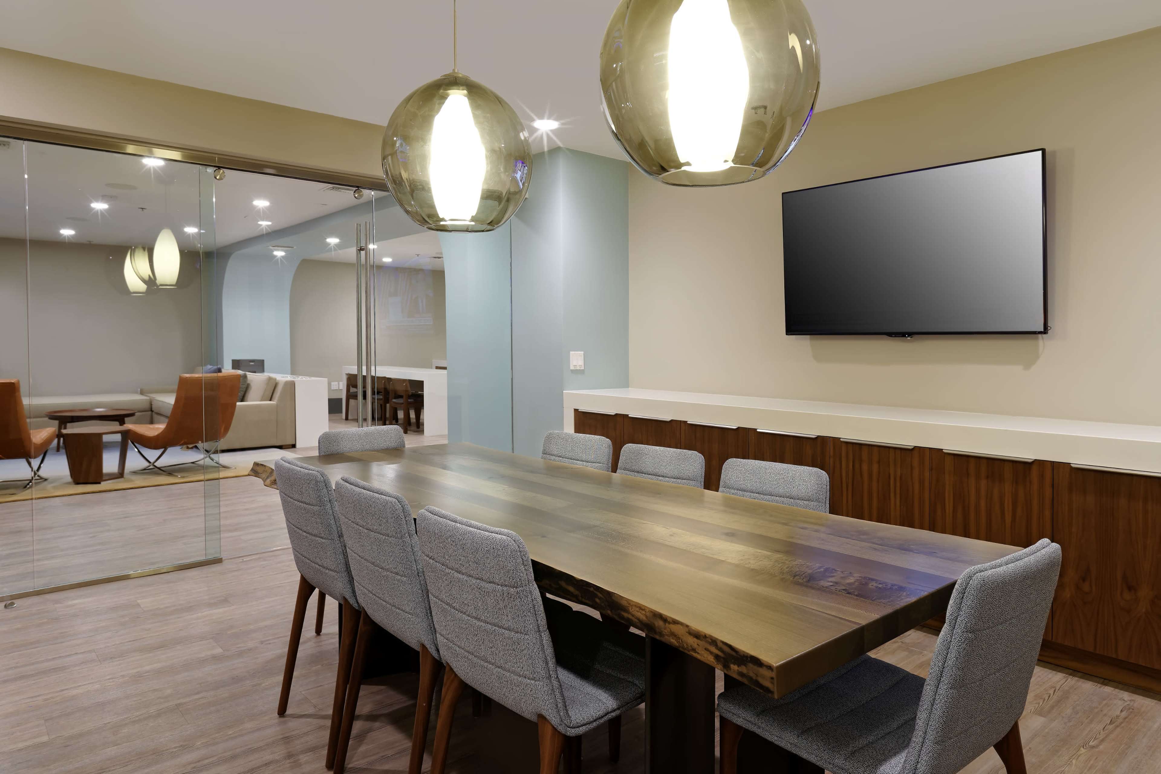 A modern conference room features a long wooden table surrounded by gray upholstered chairs, with pendant lighting overhead and a large television mounted on the wall.