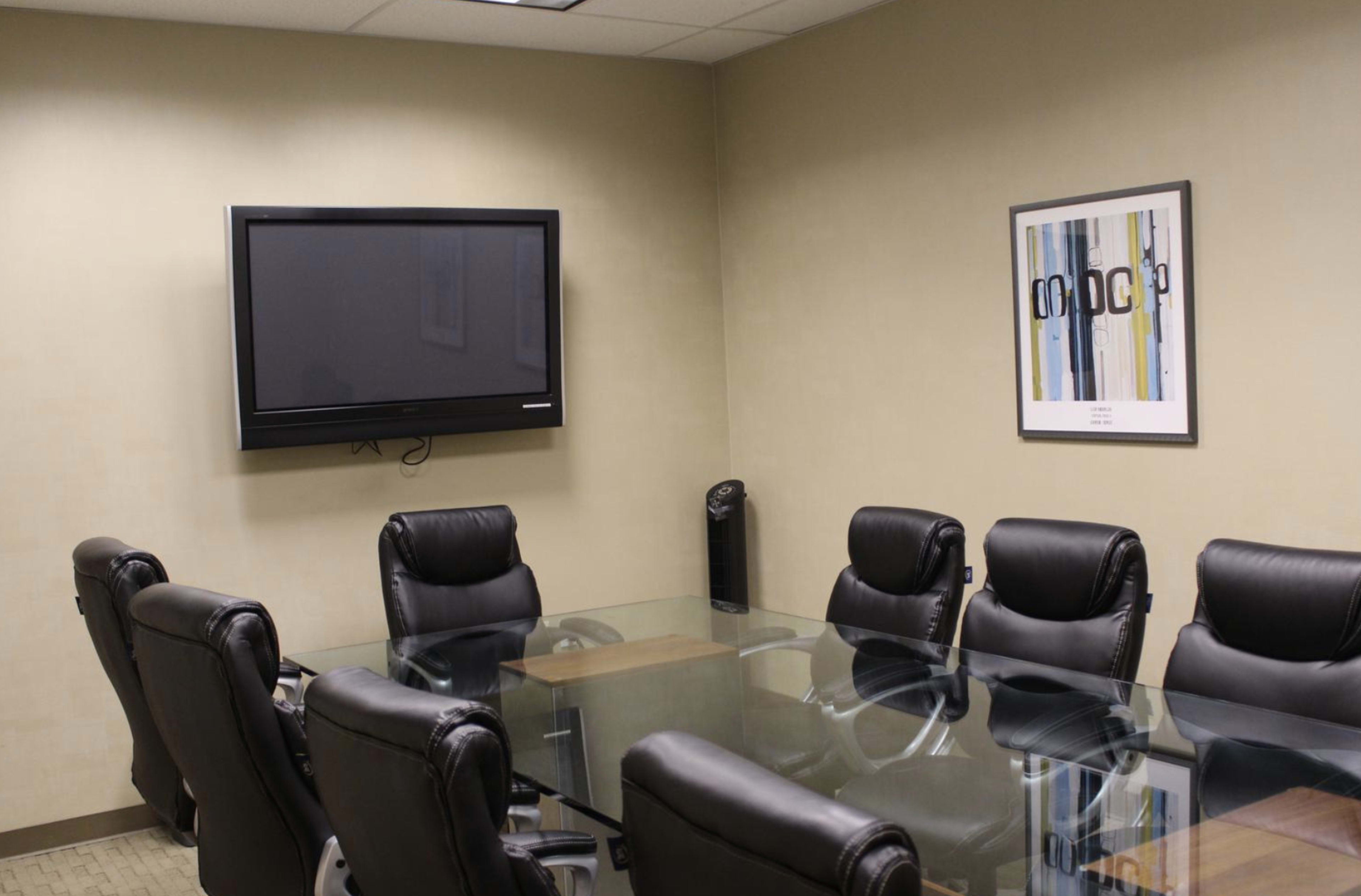 A conference room features a large glass table surrounded by black leather chairs, with a wall-mounted television and a framed abstract artwork.