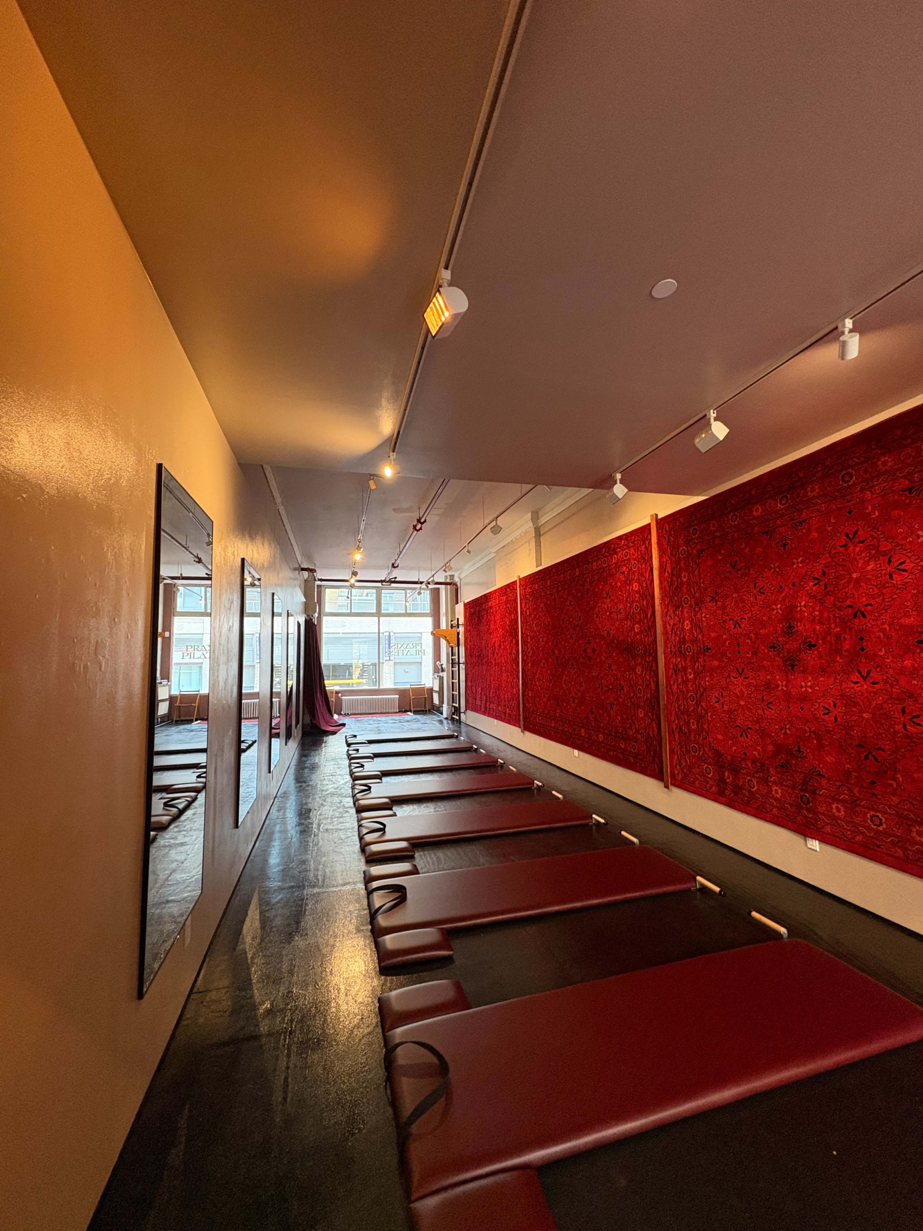 The image shows a spacious, minimalist studio with padded mats arranged in rows against a wall adorned with large, red textile panels.
