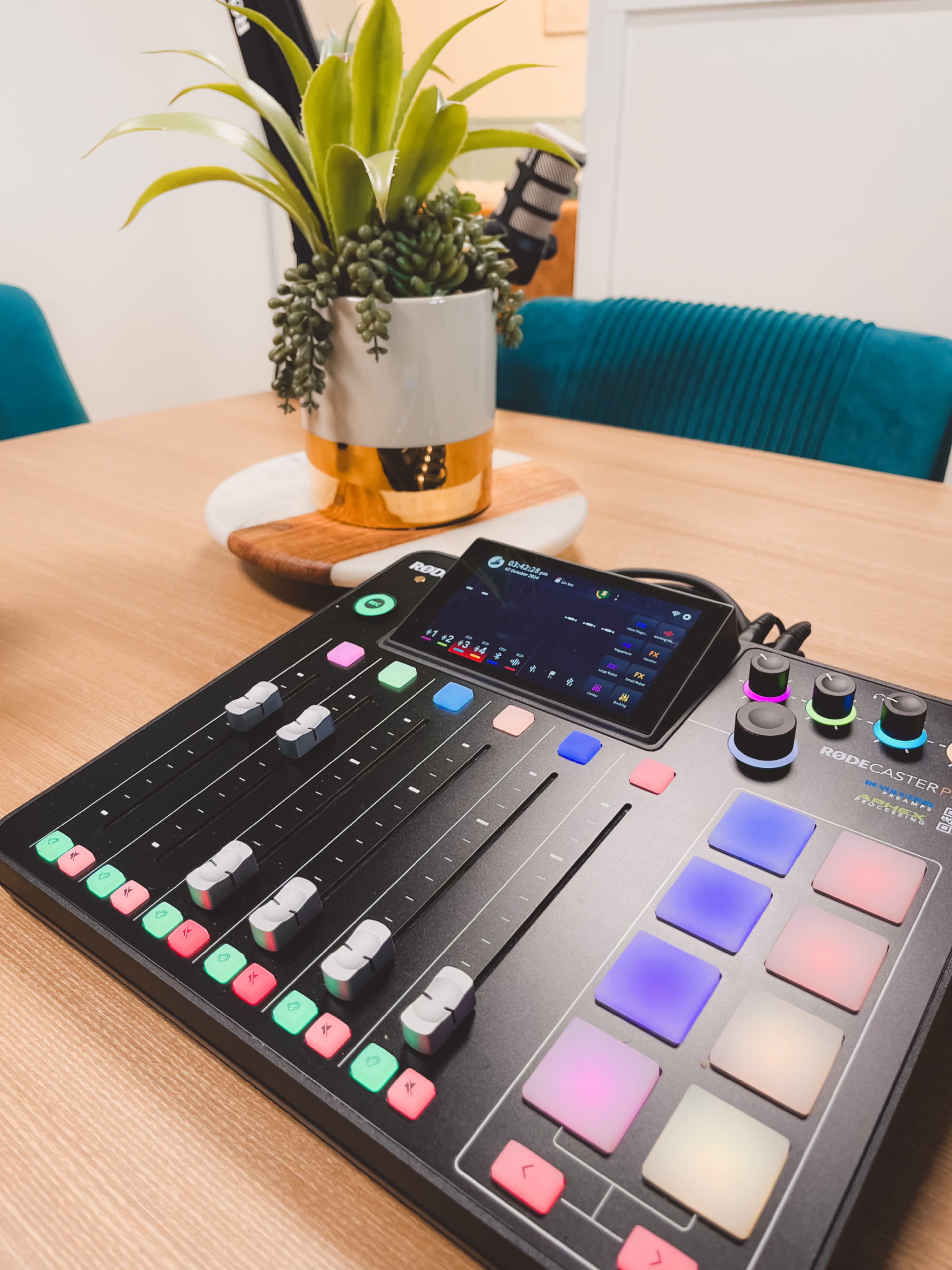 The image shows a podcasting control surface with various sliders, buttons, and a small screen on a wooden table next to a potted plant.