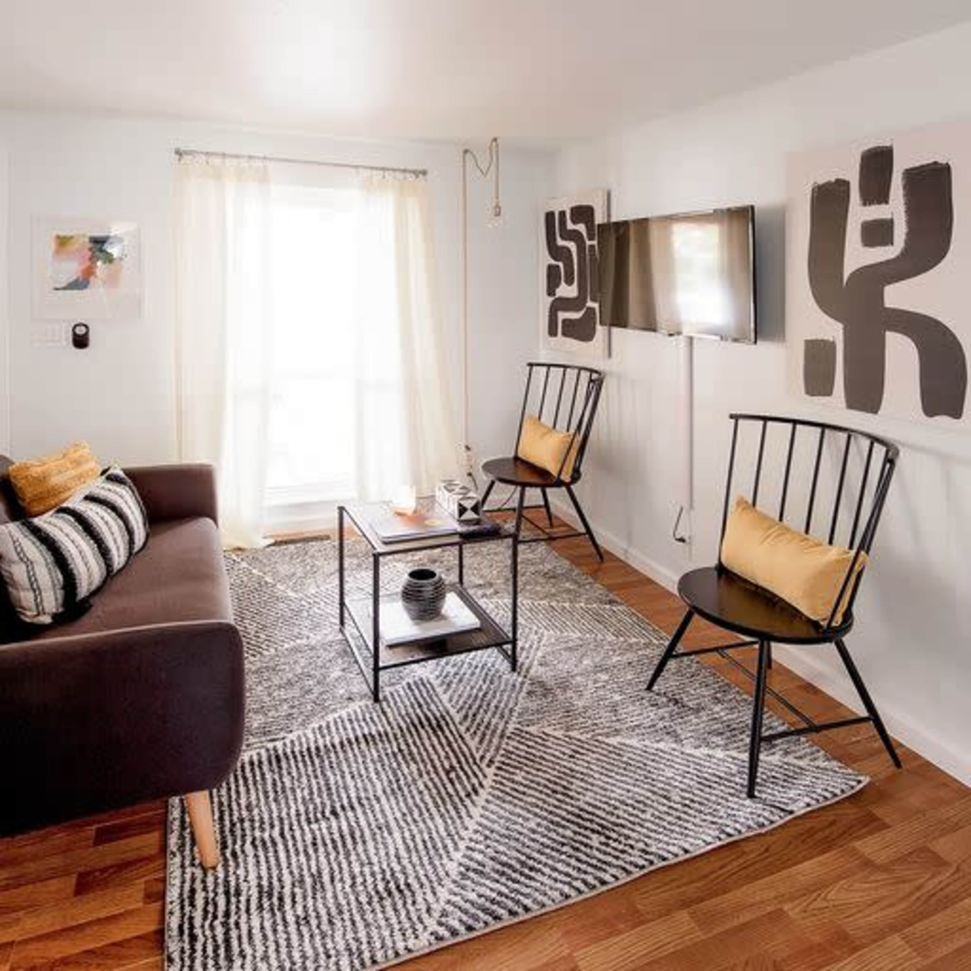 A cozy living room features a brown sofa, two black chairs, a coffee table, and a patterned area rug, all arranged around a large window.