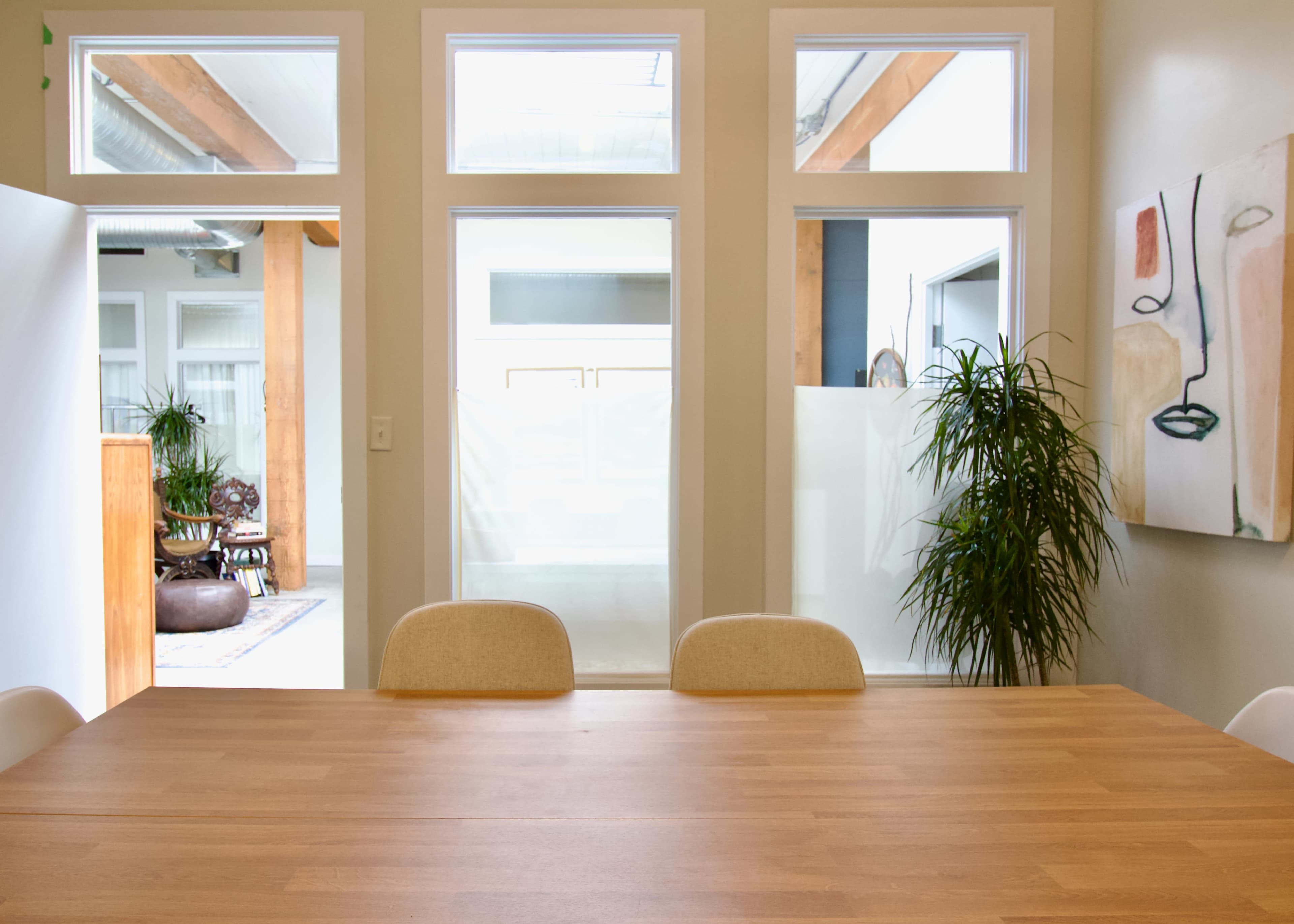 The image shows a light-filled room with a wooden table and several chairs, surrounded by large windows and a potted plant.