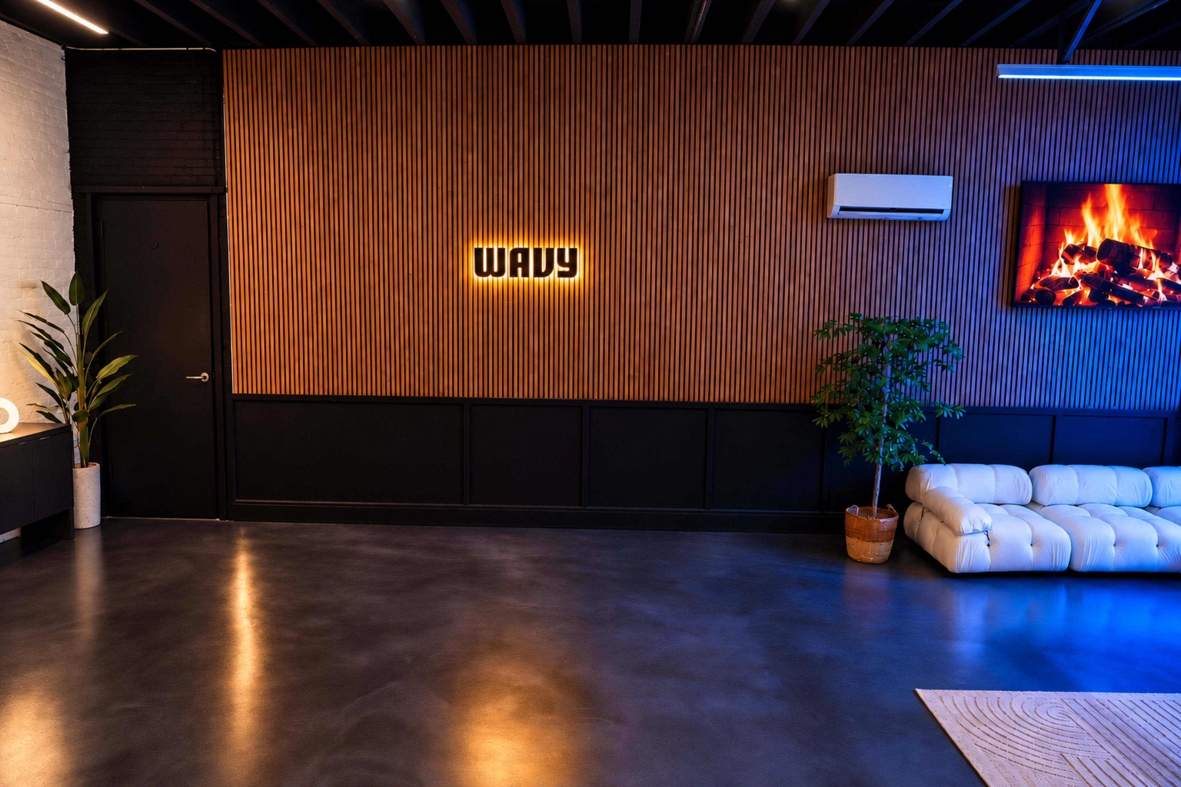The image shows a modern interior space with a textured wooden wall, a white sofa, a potted plant, and a wall-mounted television displaying a fireplace.