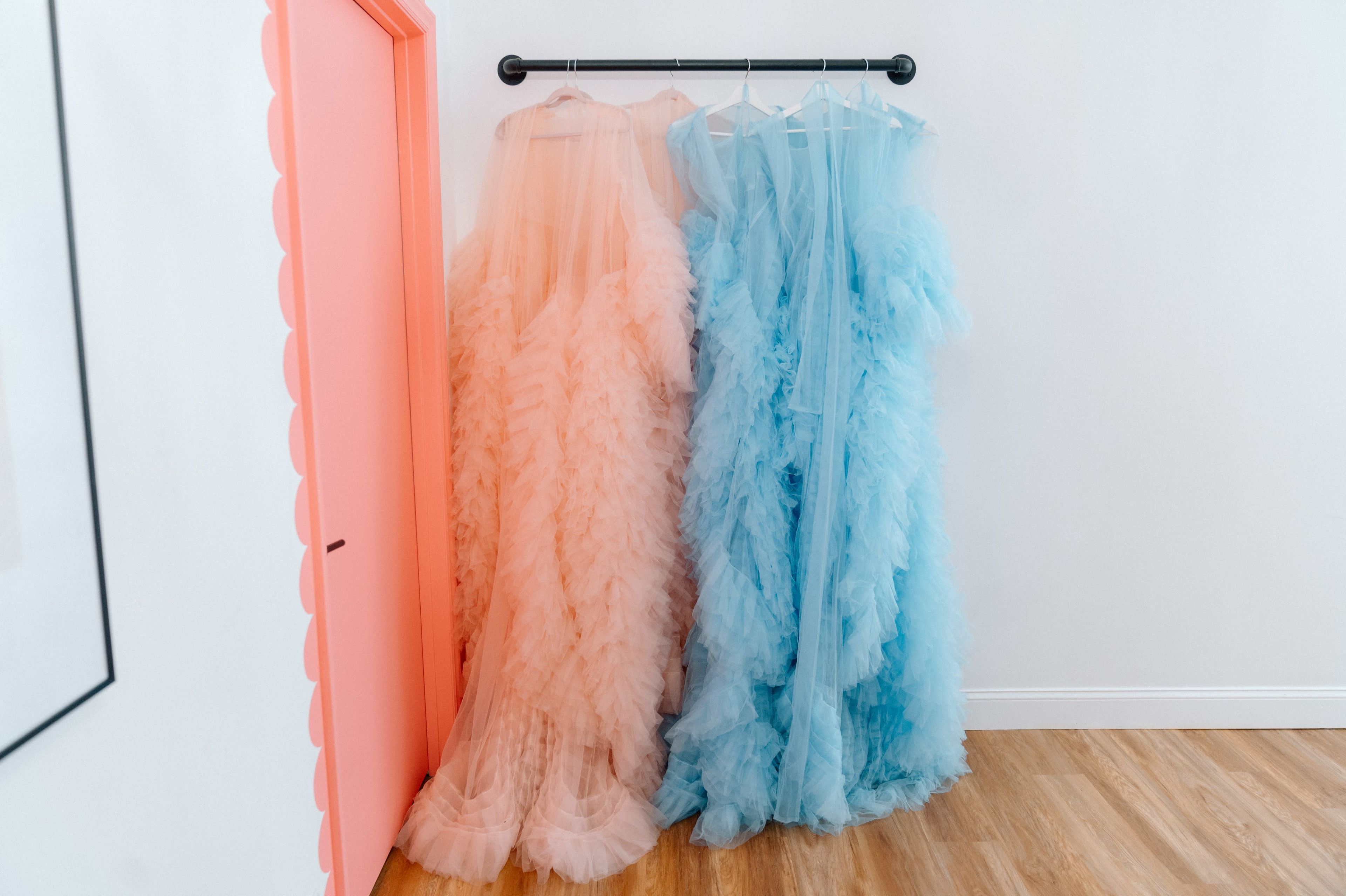 The image features a rack holding two fluffy, oversized dresses, one in soft peach and the other in light blue, against a white wall with a coral door.