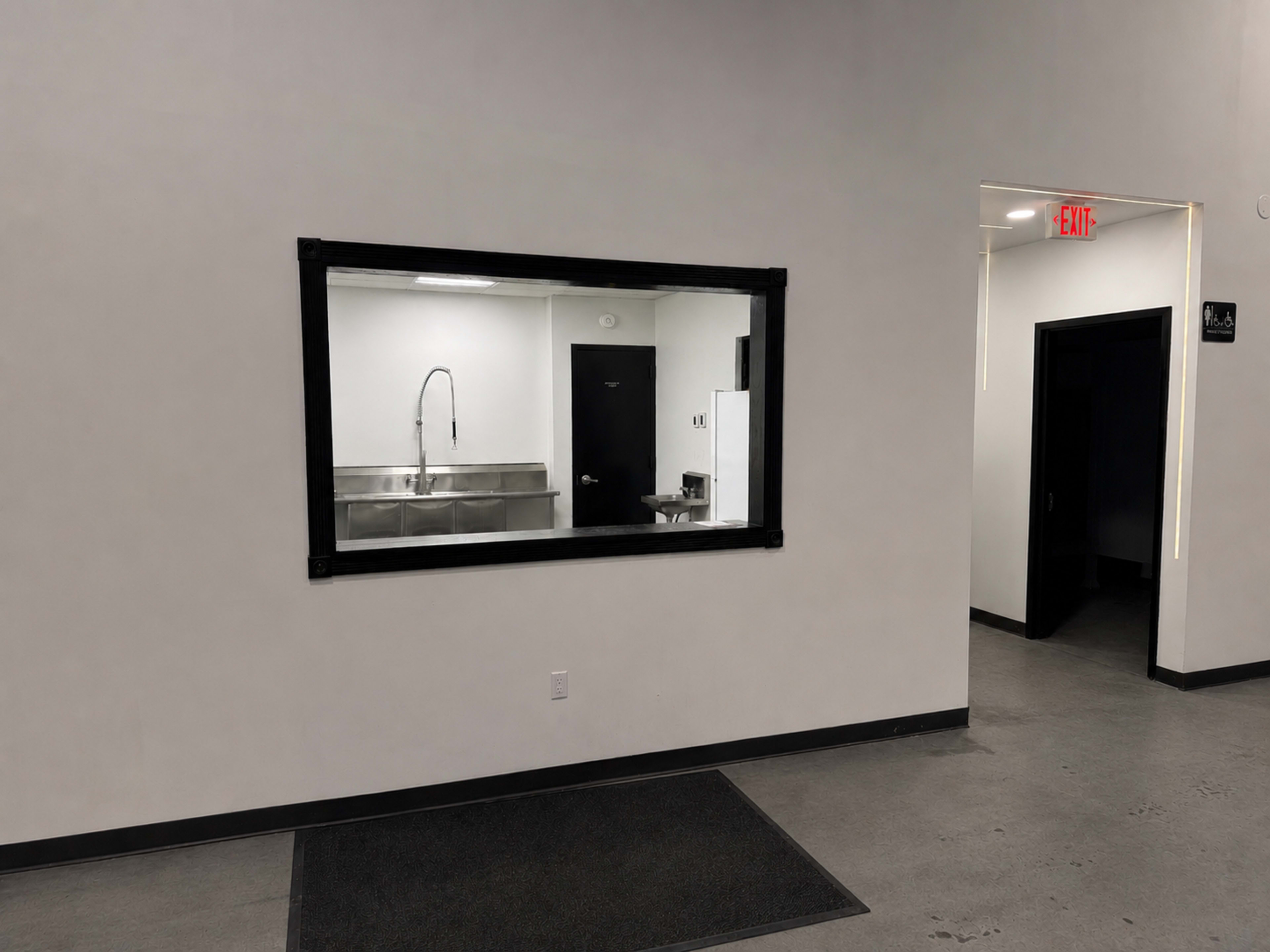 The image shows a clean, minimalist corridor with a large window looking into a room featuring a stainless steel sink and fixtures.