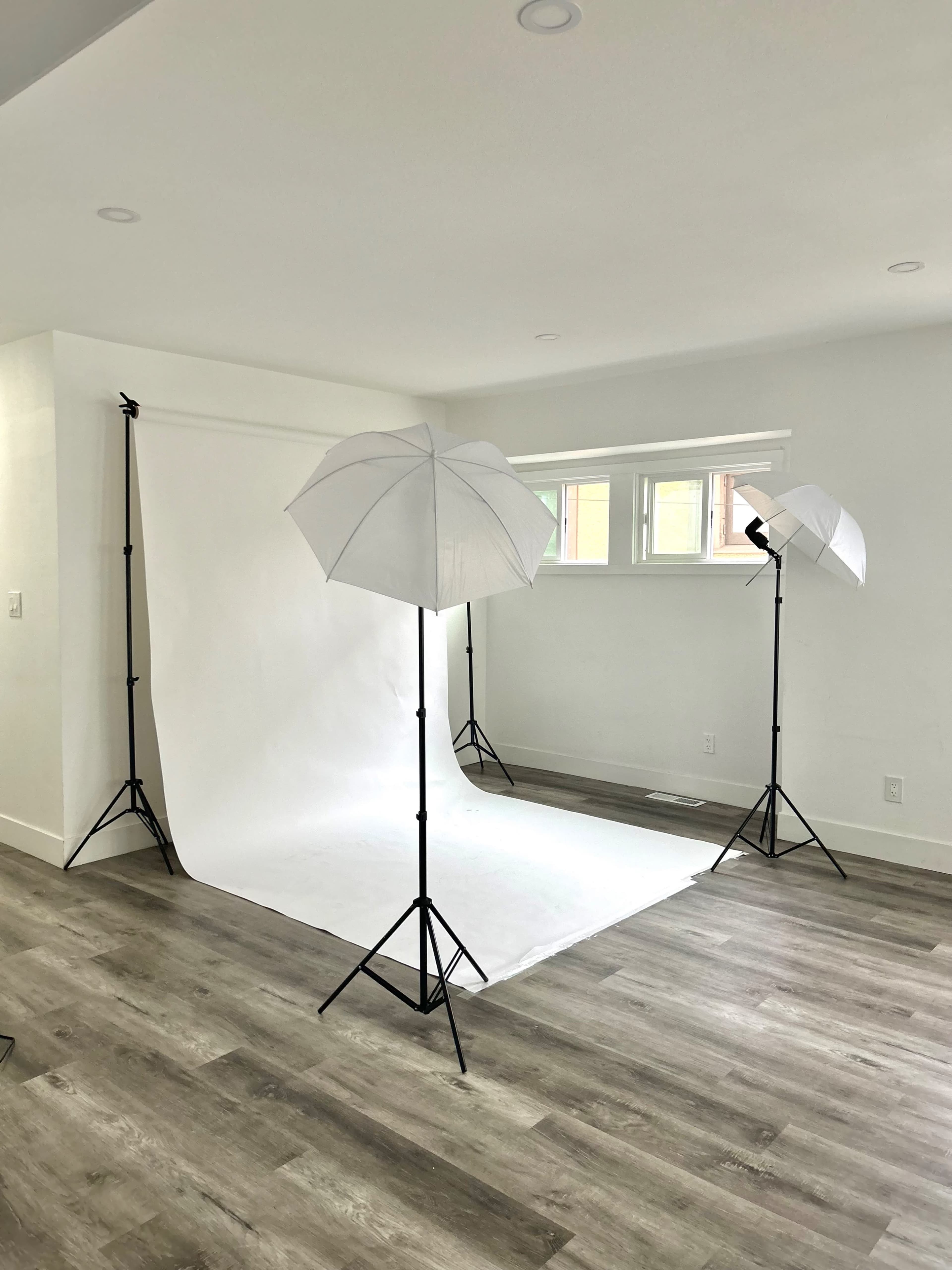 The image shows a photography studio with a white backdrop, lighting umbrellas on stands, and light-colored wooden flooring.