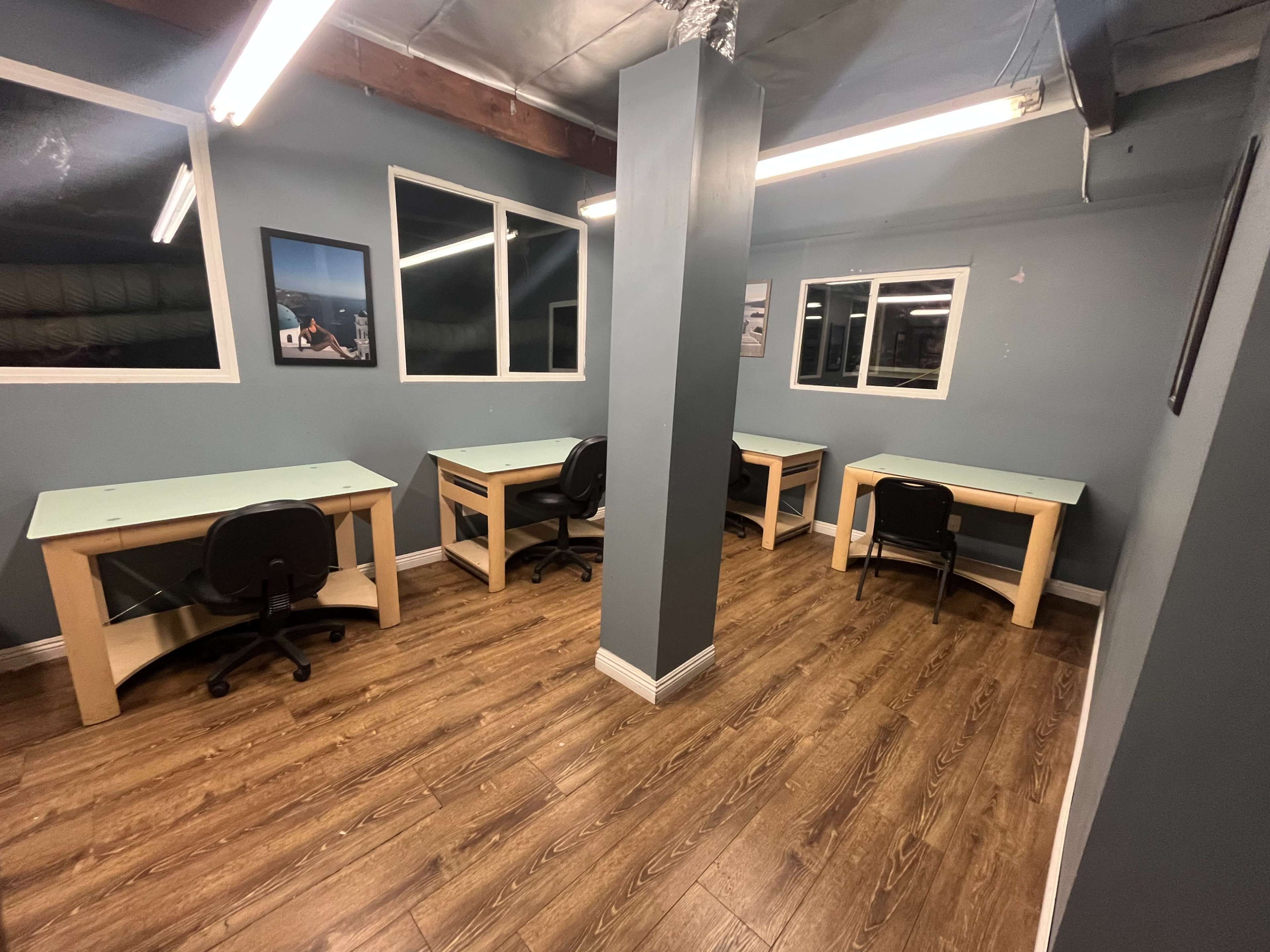 The image shows a small office space with four desks and black chairs arranged on a wooden floor, surrounded by blue walls and large windows.