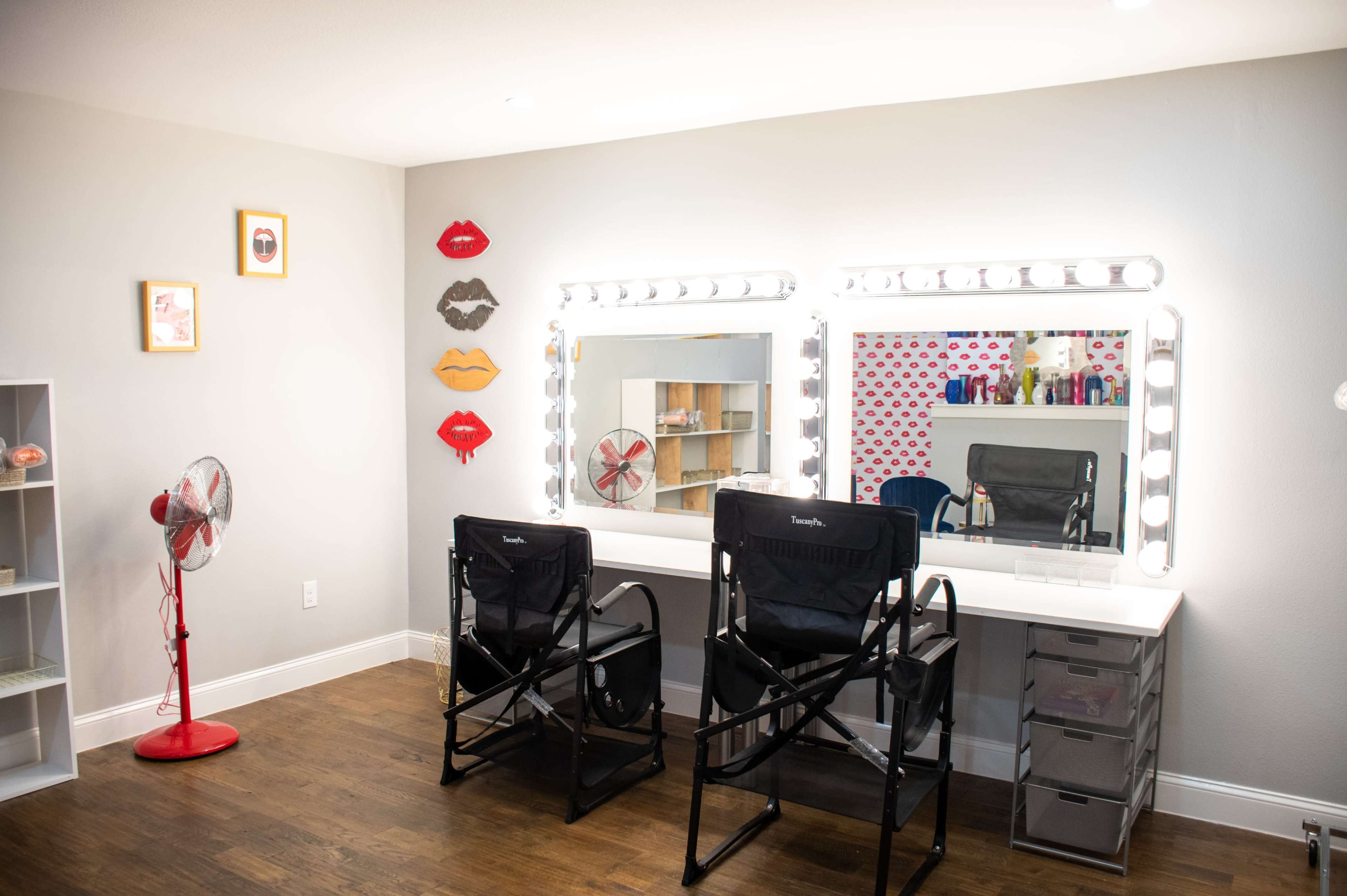 A brightly lit makeup room features two director's chairs in front of large illuminated mirrors, with a fan and decorative wall art nearby.