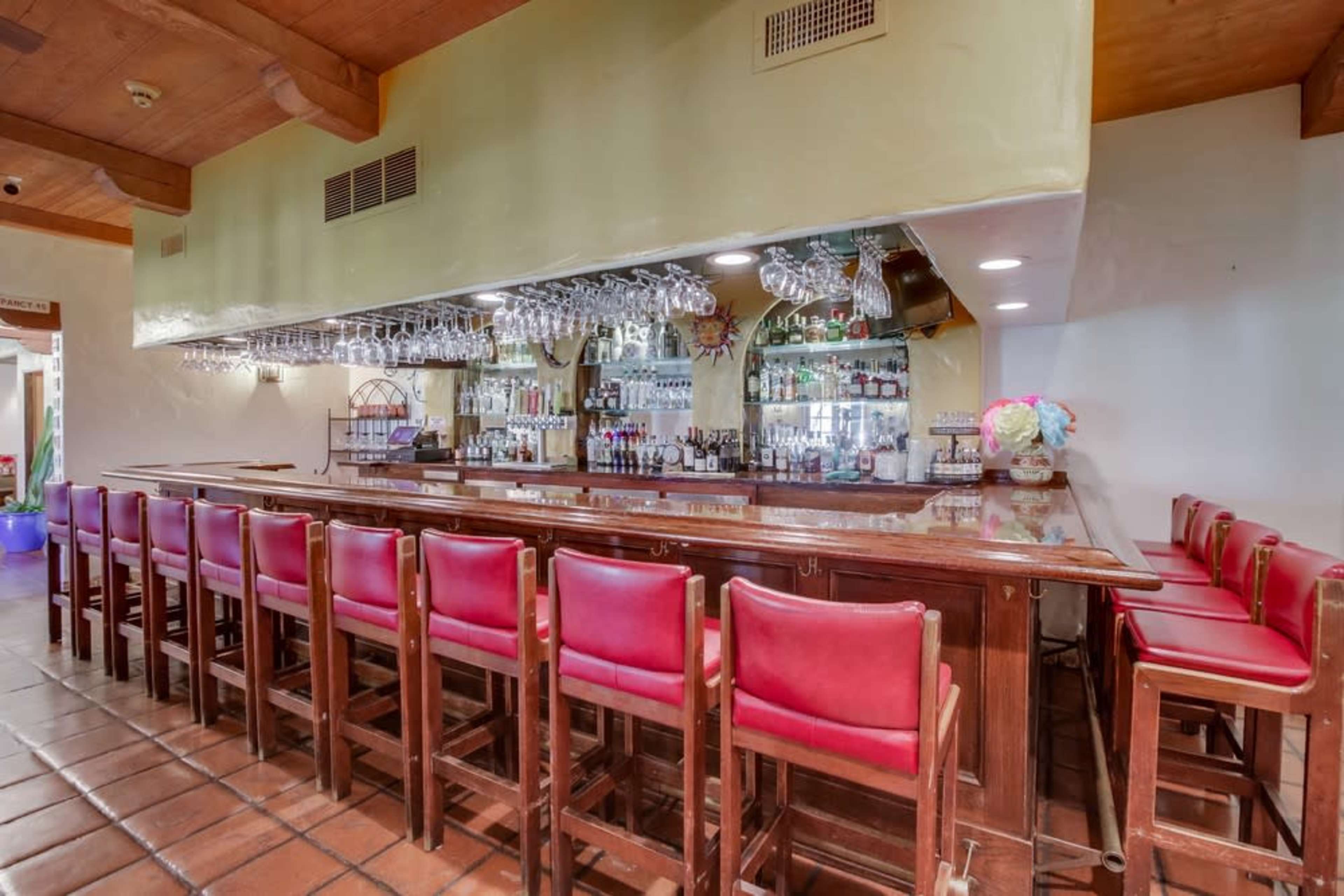 The image shows a bar area with a long wooden counter, red upholstered stools, and shelves lined with various bottles and glasses.