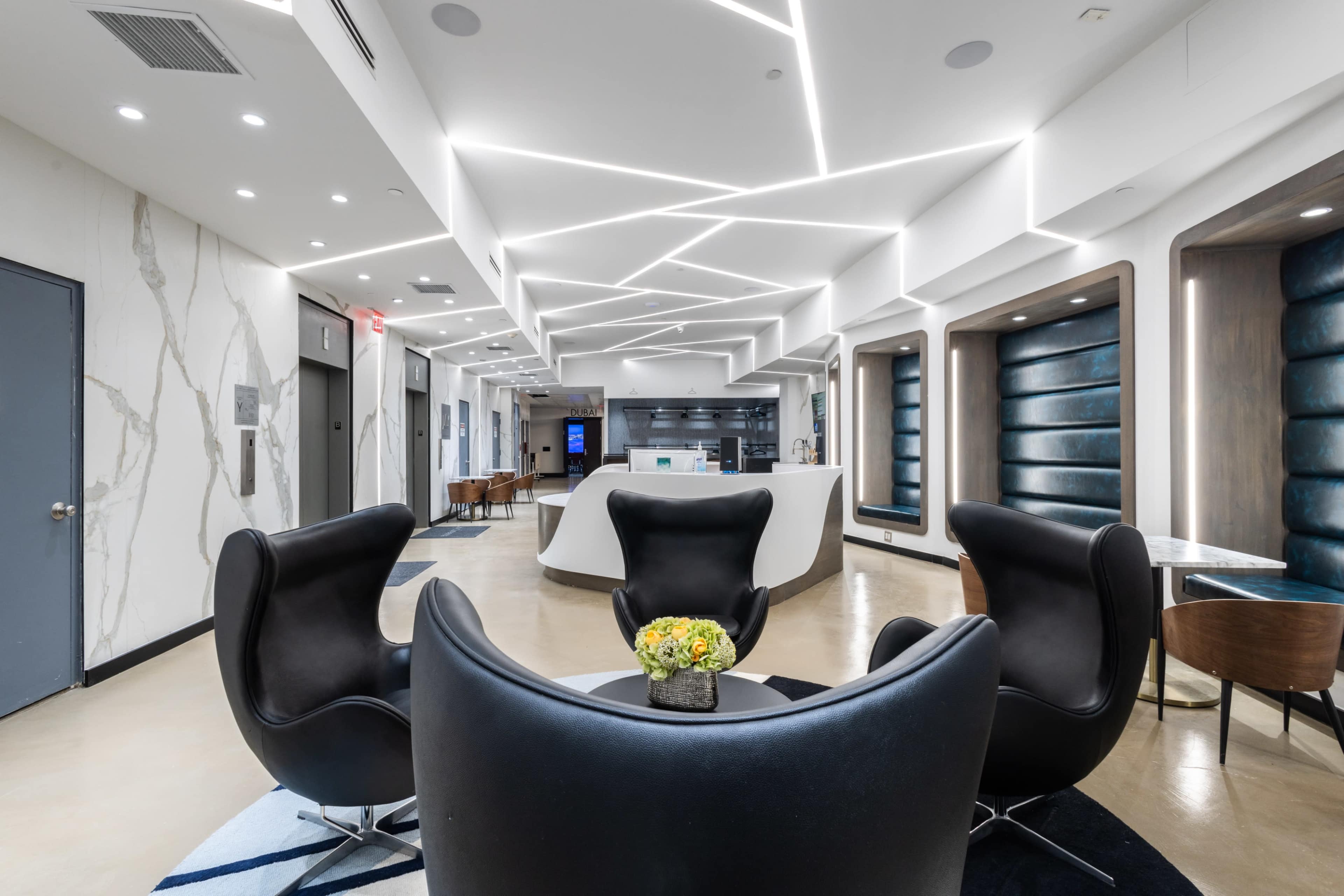 The image shows a modern lobby featuring sleek furniture, geometric ceiling lights, and a reception area.