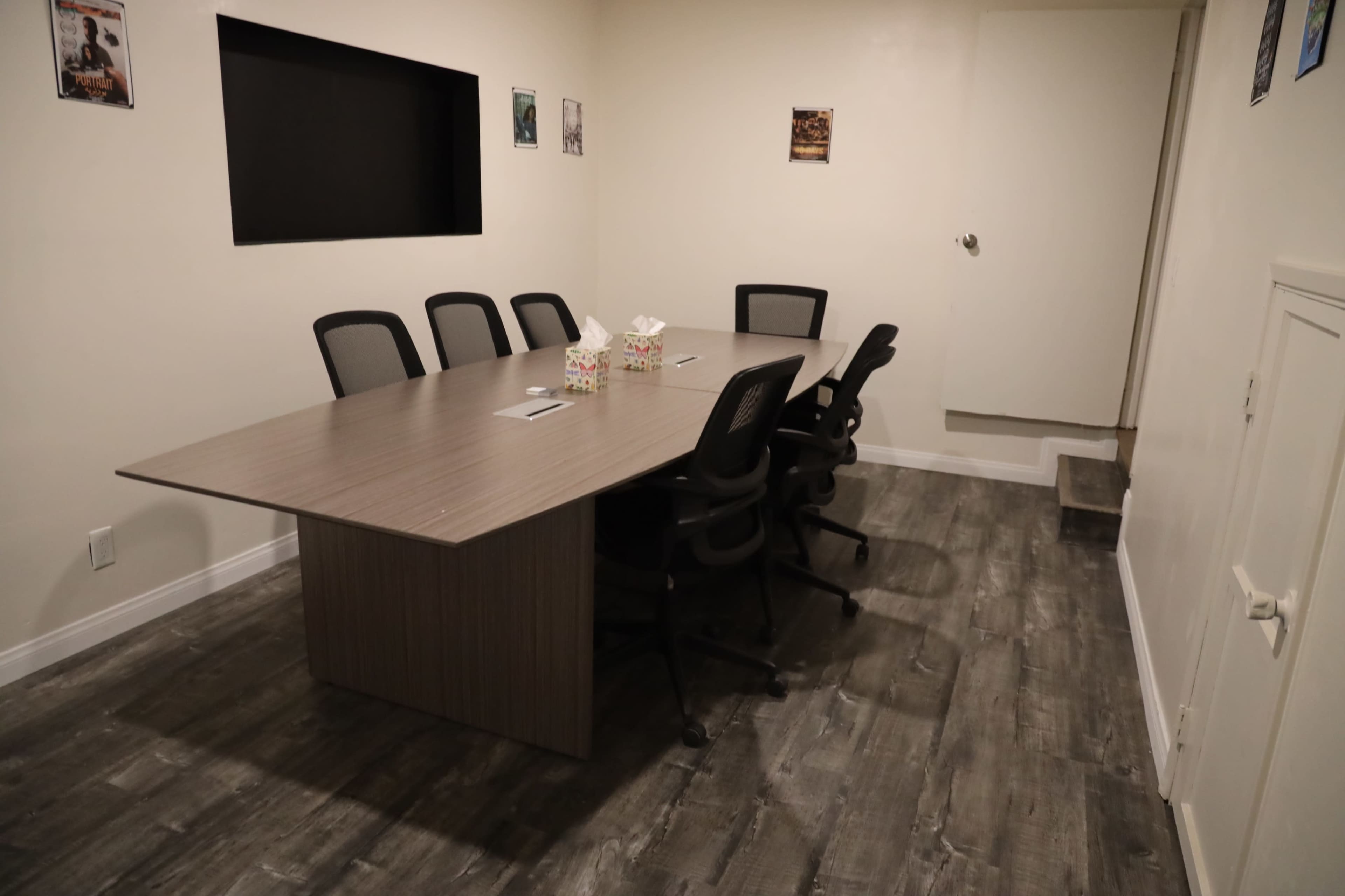 A conference room features a large table surrounded by six black chairs, with a wall-mounted screen and a door leading to another area.