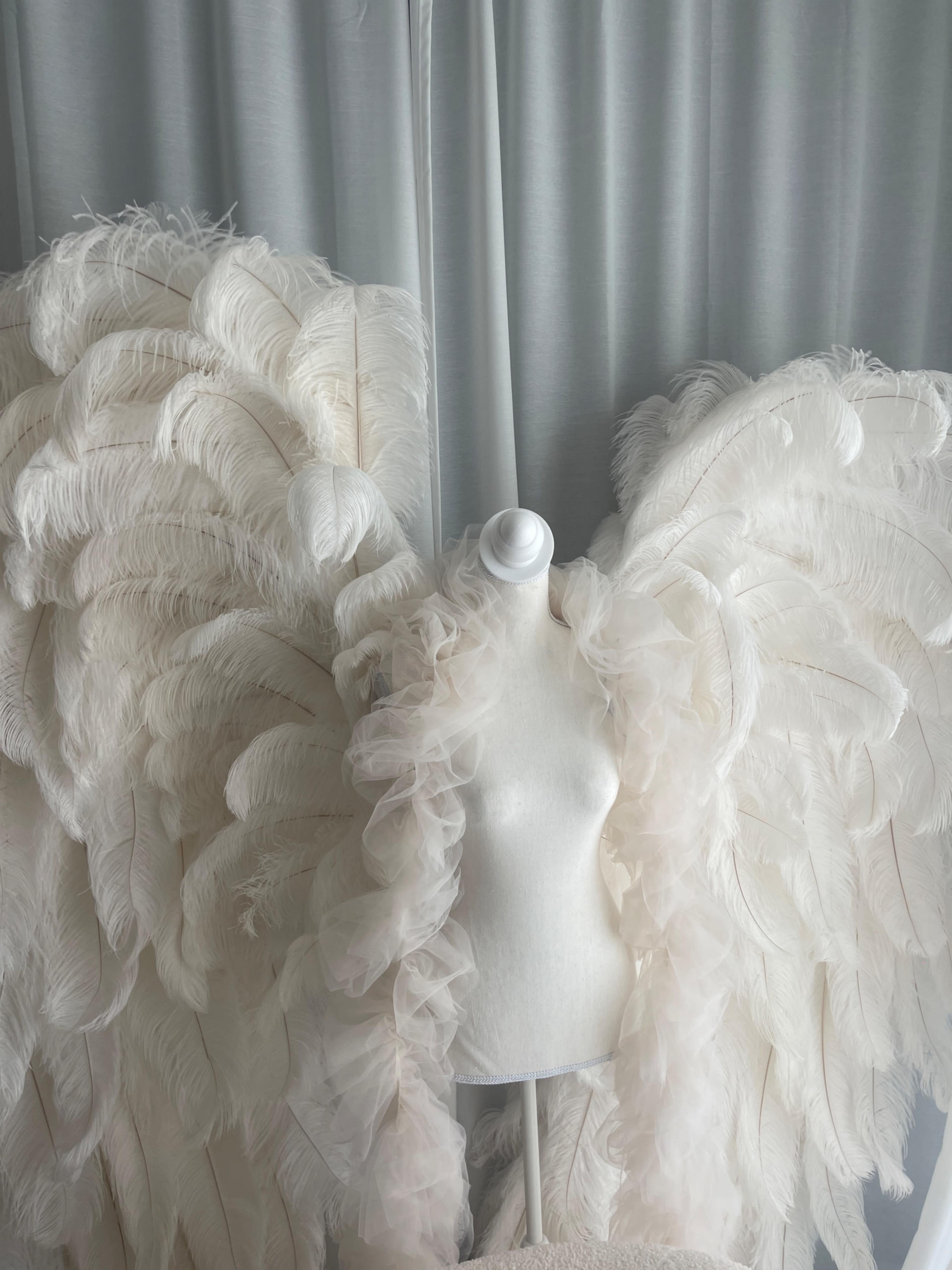 The image shows a mannequin adorned with large, white feathered wings positioned against a backdrop of light-colored fabric.
