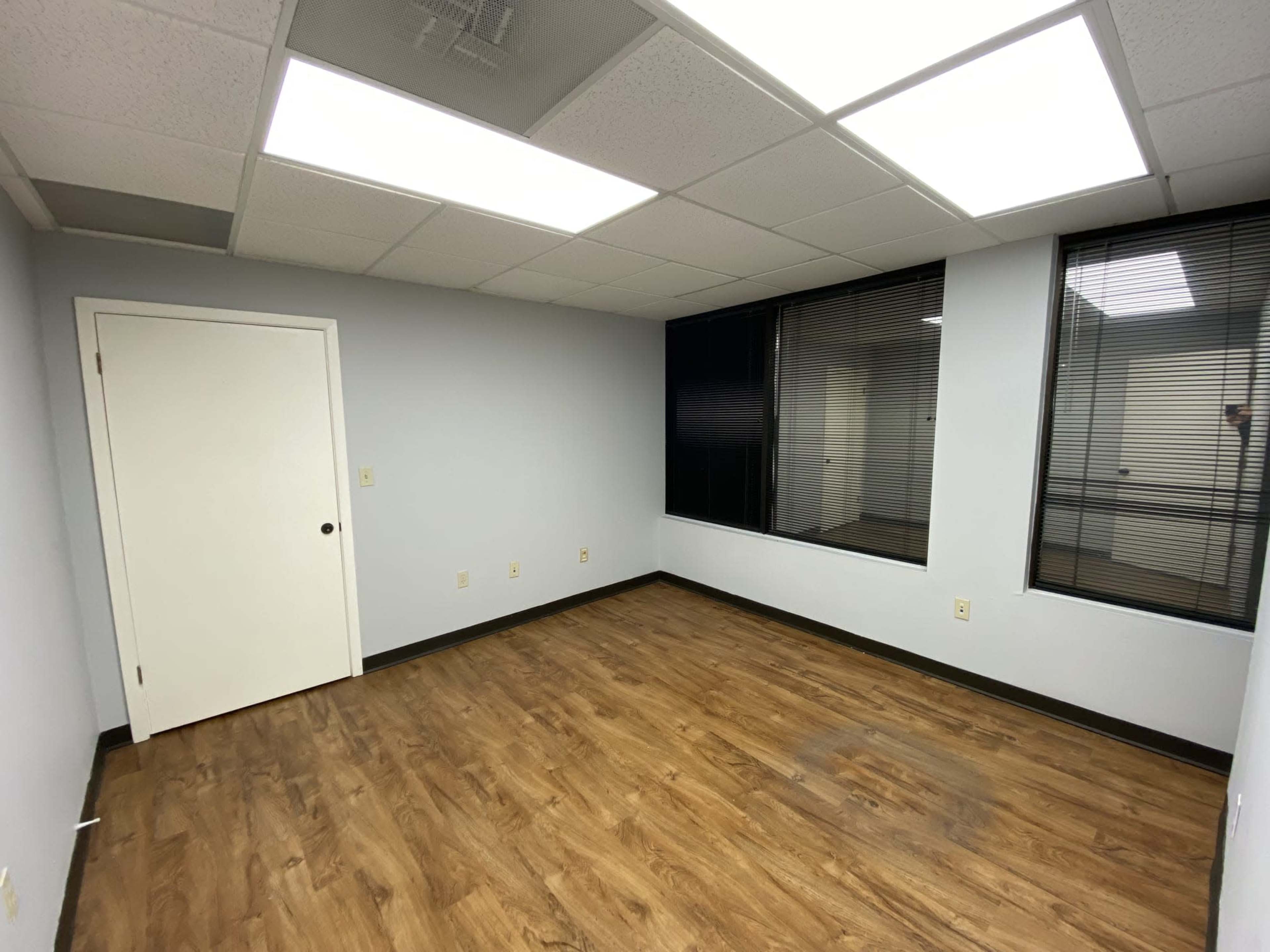 The image shows an empty office room with light-colored walls, a single door, and large windows featuring blinds.