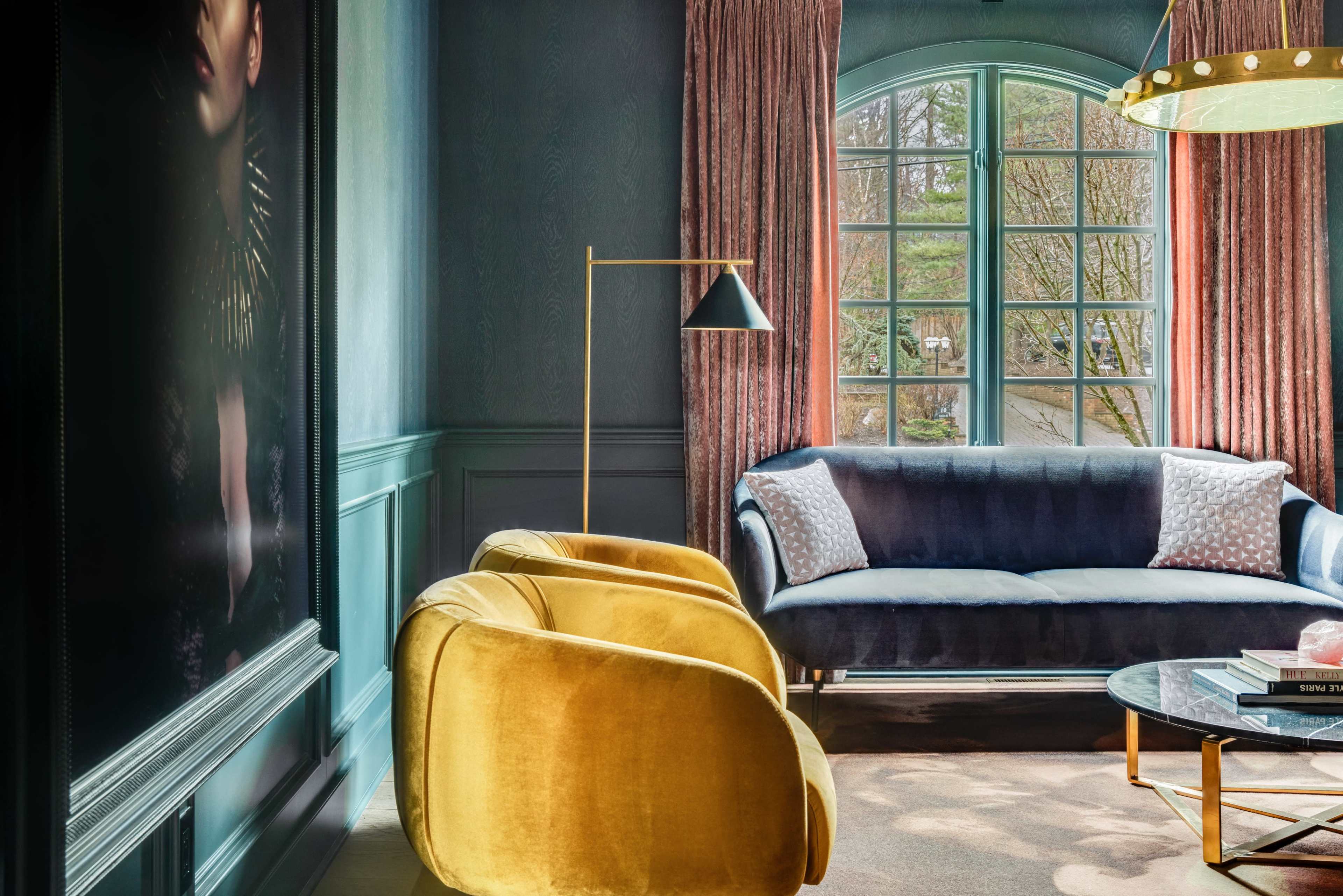 The image shows a stylish living room featuring a blue sofa, yellow armchairs, large windows with sheer curtains, and a round glass coffee table.