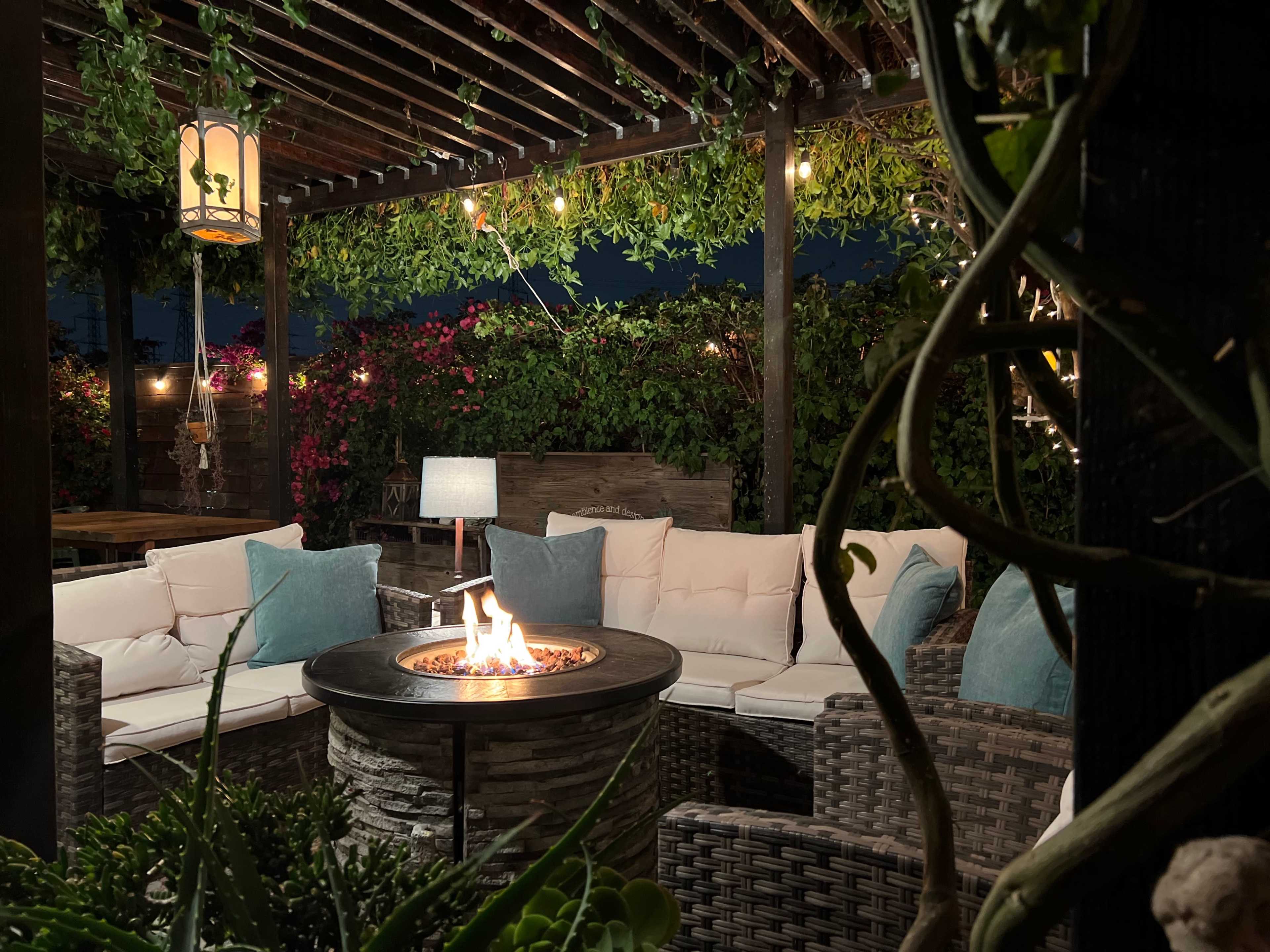 A cozy outdoor seating area features a circular fire pit surrounded by cushioned furniture, with lush greenery and string lights overhead.