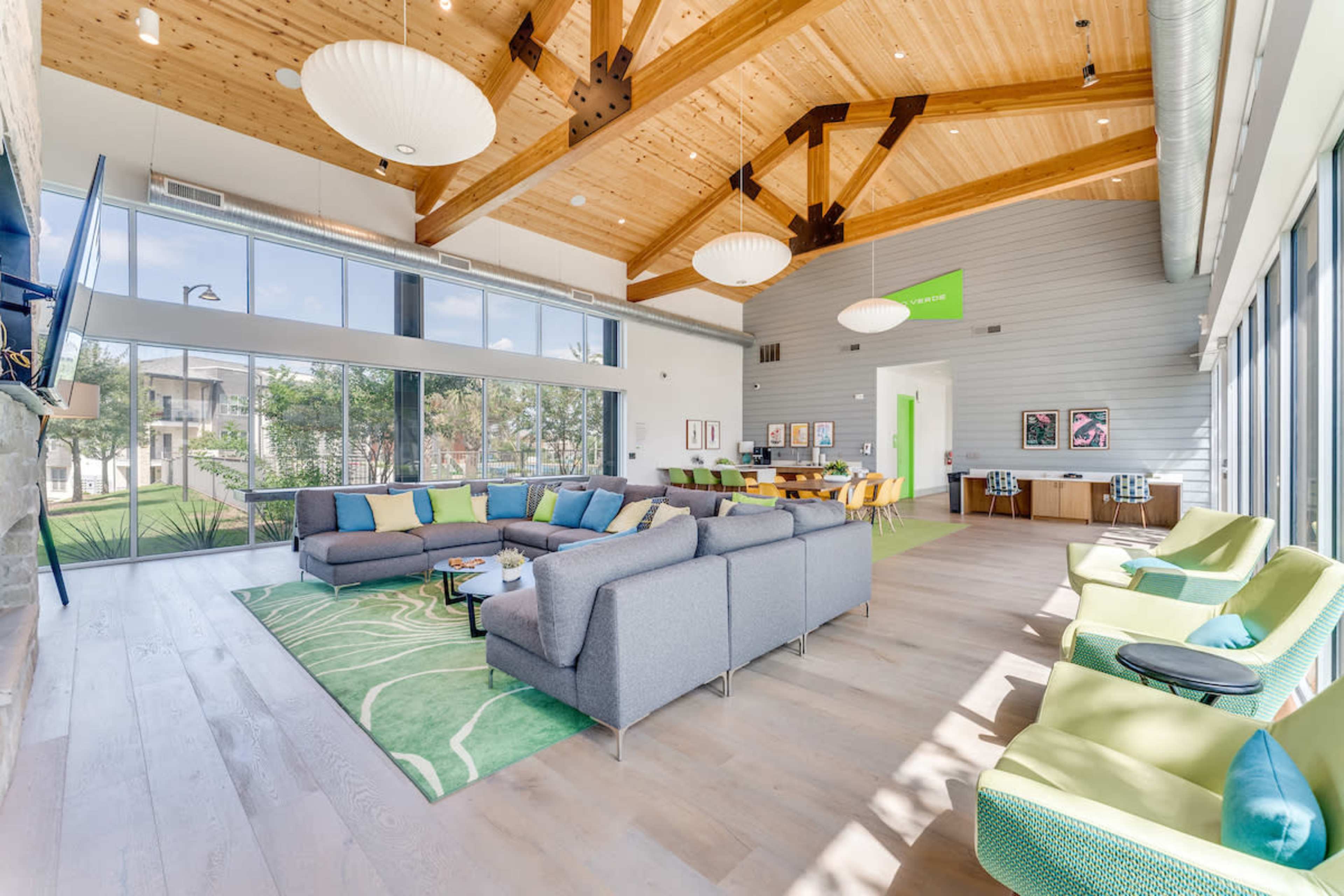 A spacious modern lounge with high wooden ceilings, large windows, and a mix of gray and colorful furniture arranged around a green area rug.