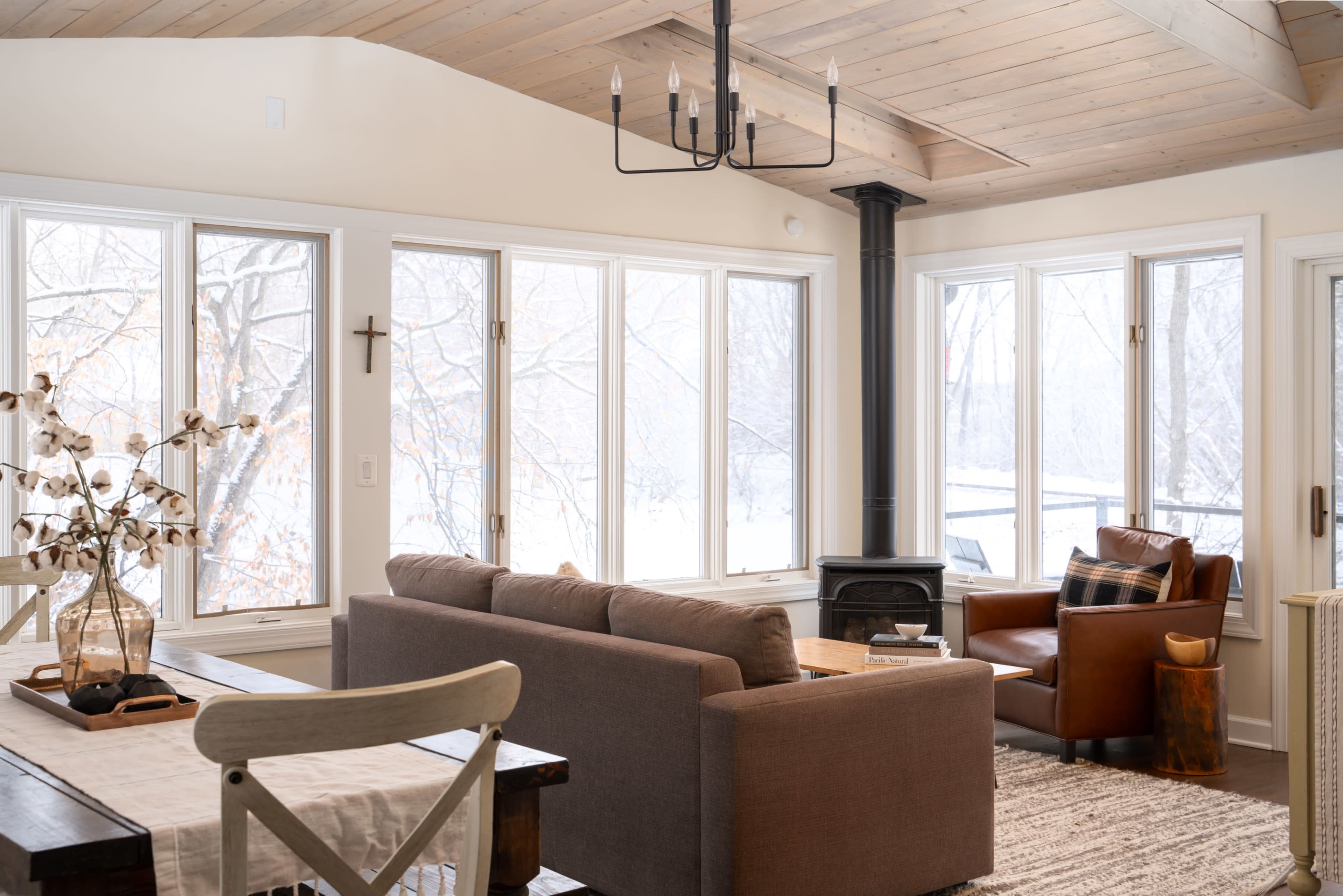 A cozy living room with large windows overlooking a snowy landscape, featuring a sofa, a leather armchair, and a wood-burning stove.