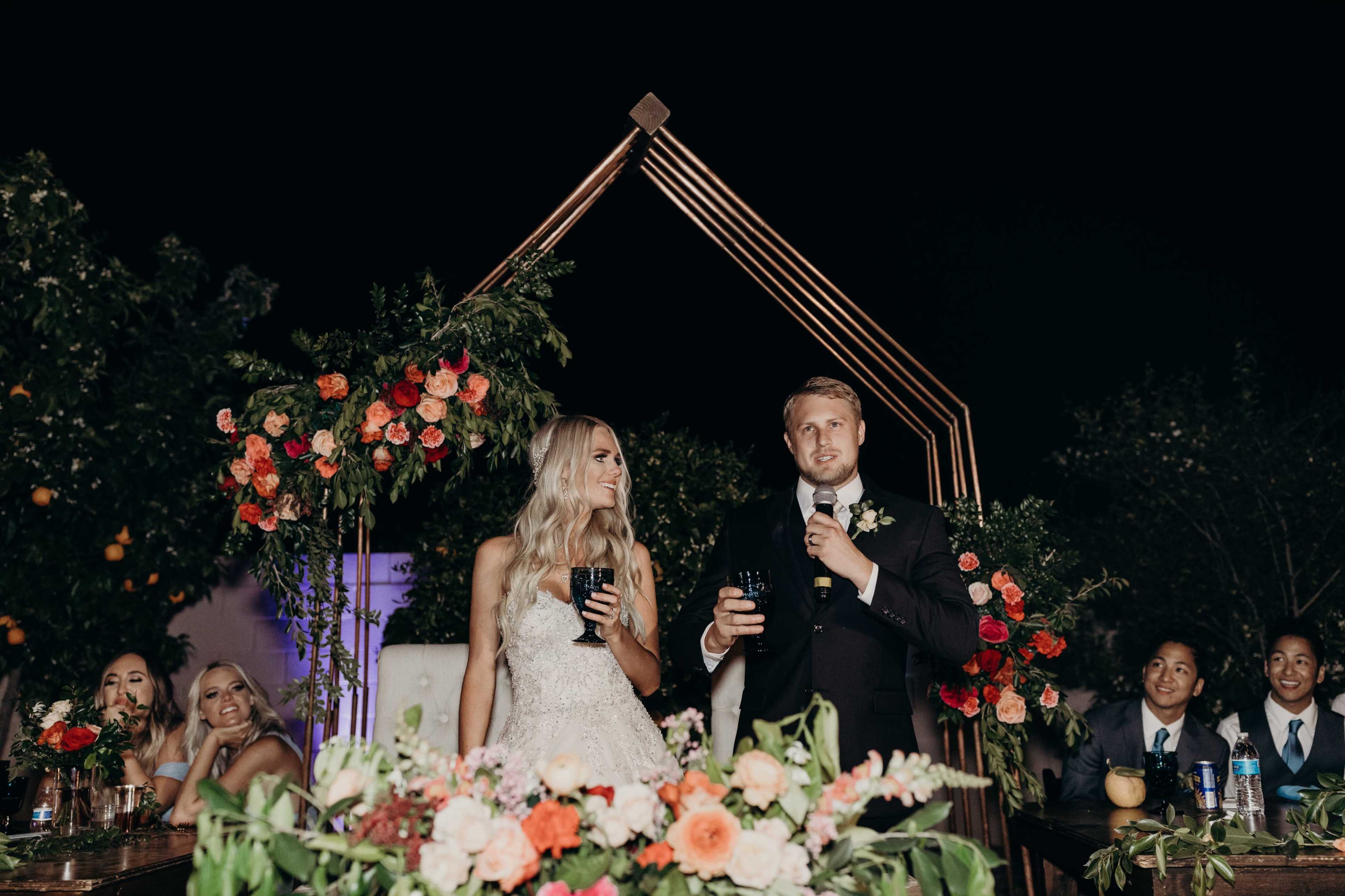 A bride and groom stand at a decorated table, speaking to guests during their wedding reception under a night sky adorned with floral arrangements.