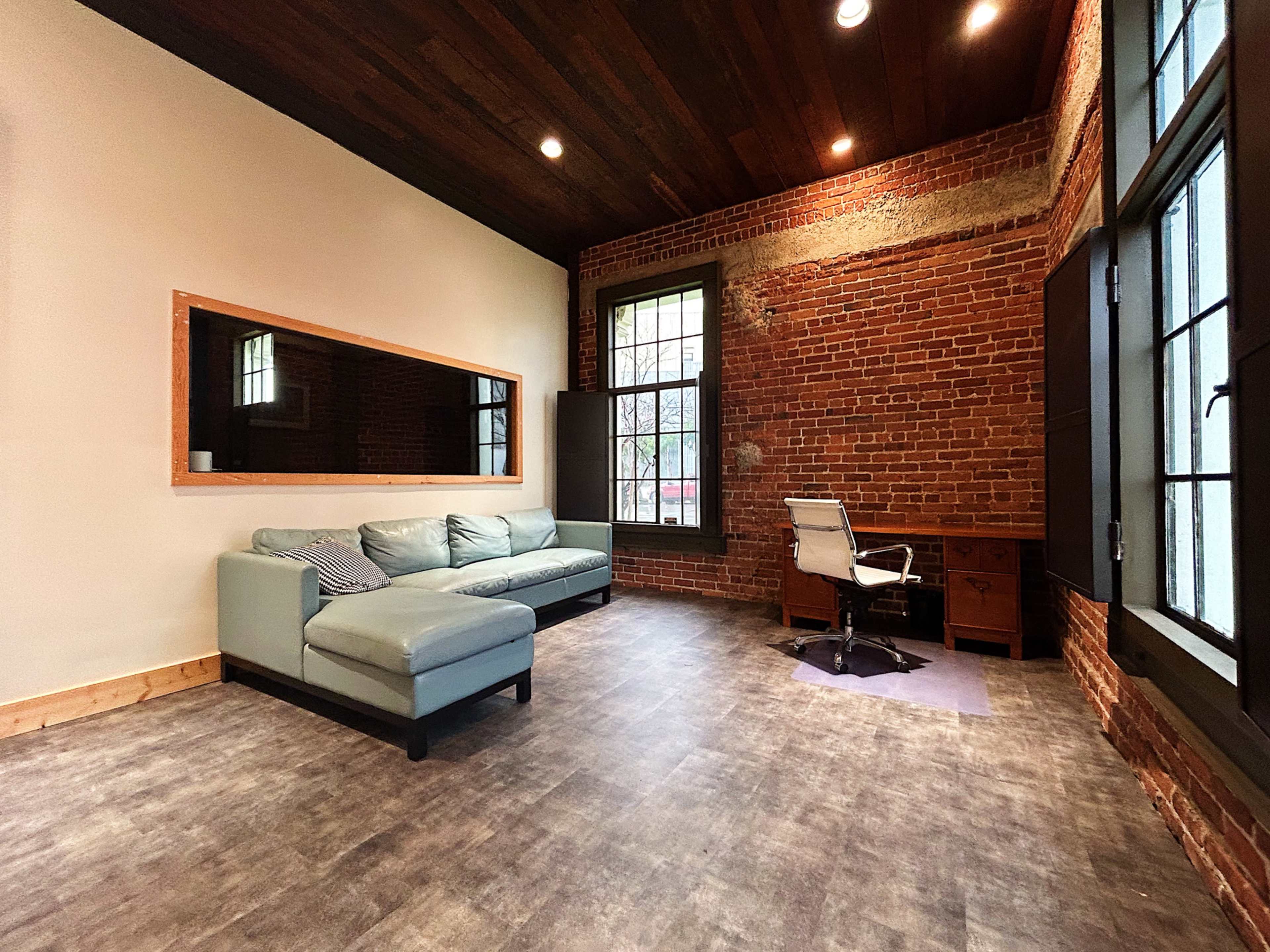 A modern office space featuring a light-colored sectional couch, a desk with a chair, and exposed brick walls.