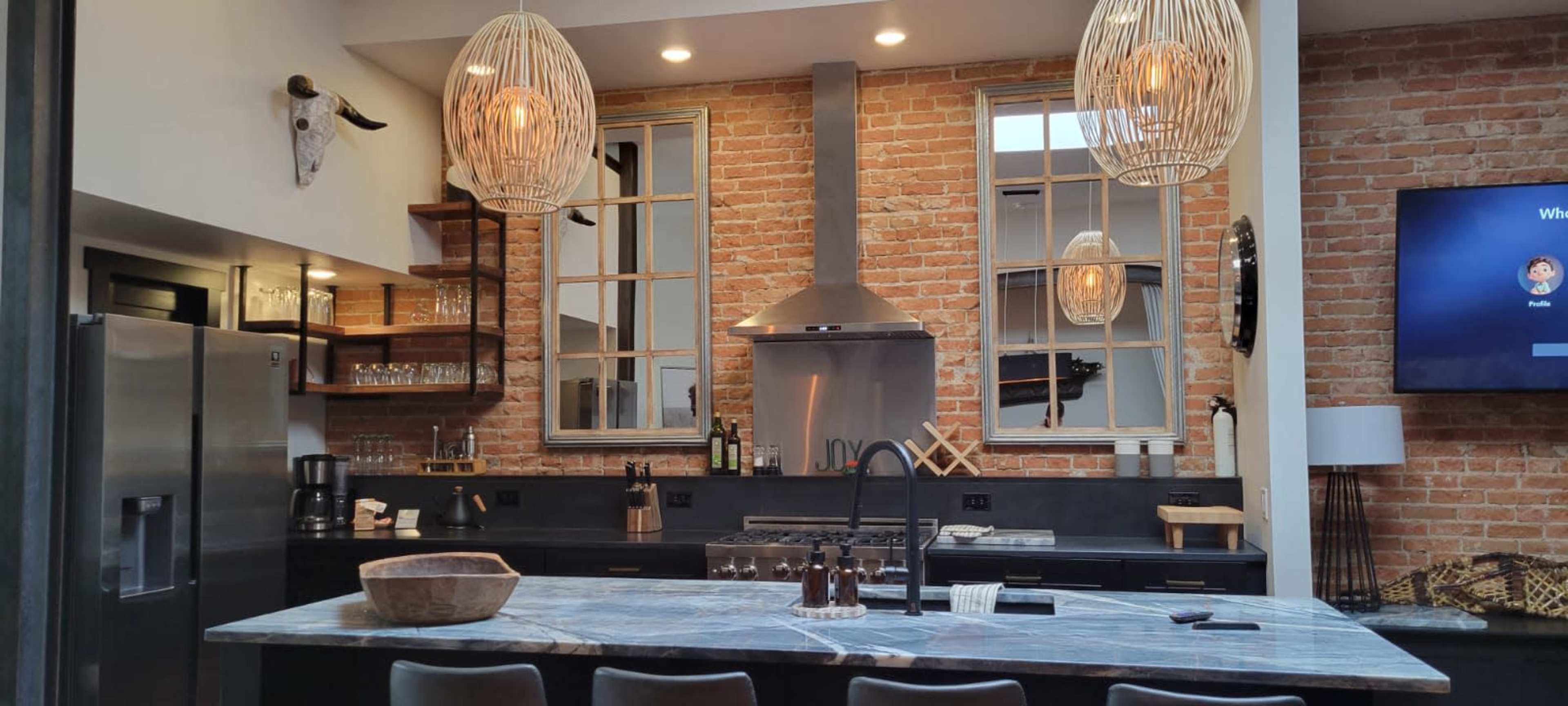 A modern kitchen features exposed brick walls, a large marble countertop, and overhead pendant lighting.