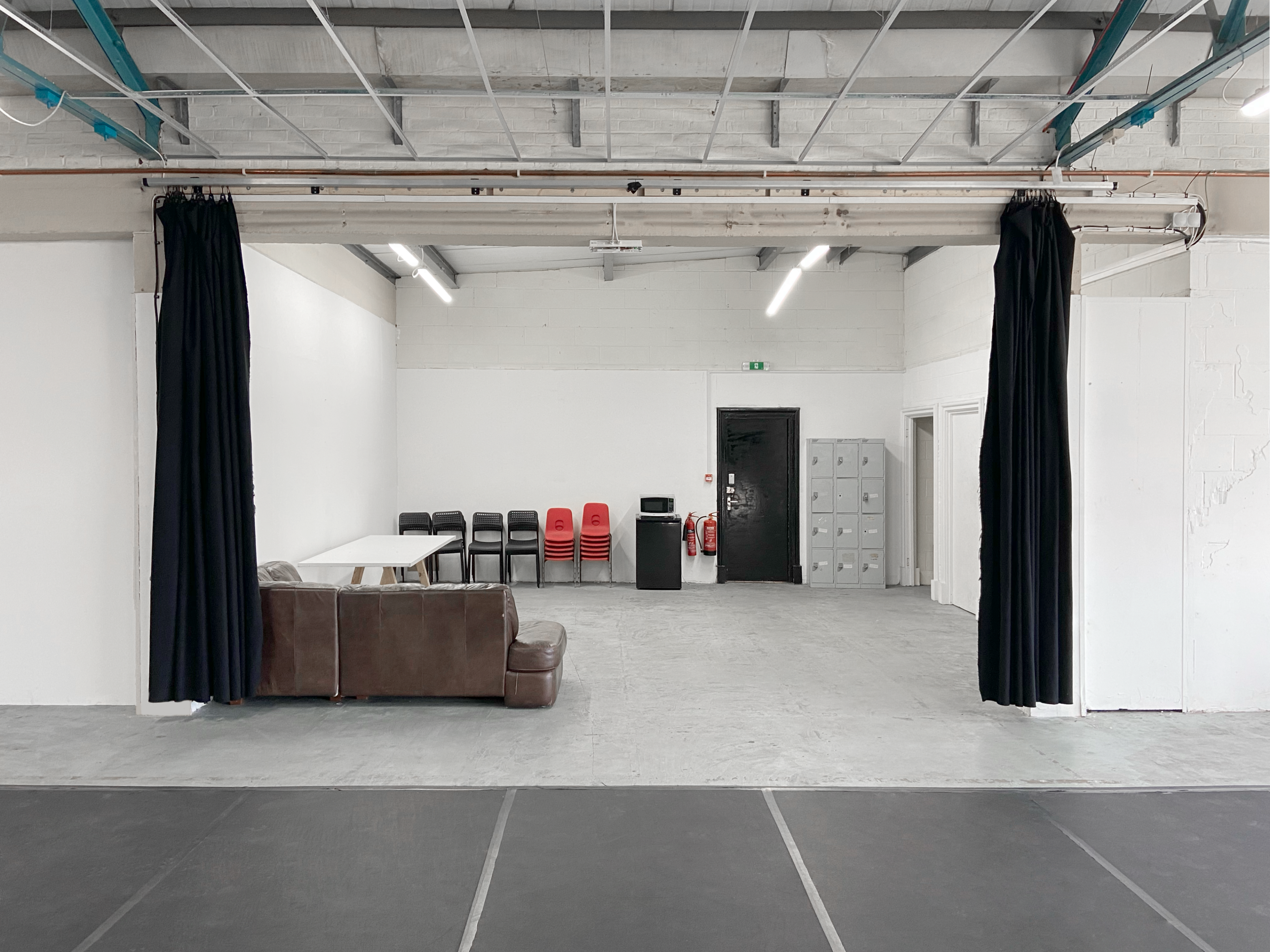 A spacious room features a seating area with a brown couch, a stack of red chairs, a table, and lockers against the walls, separated by black curtains.