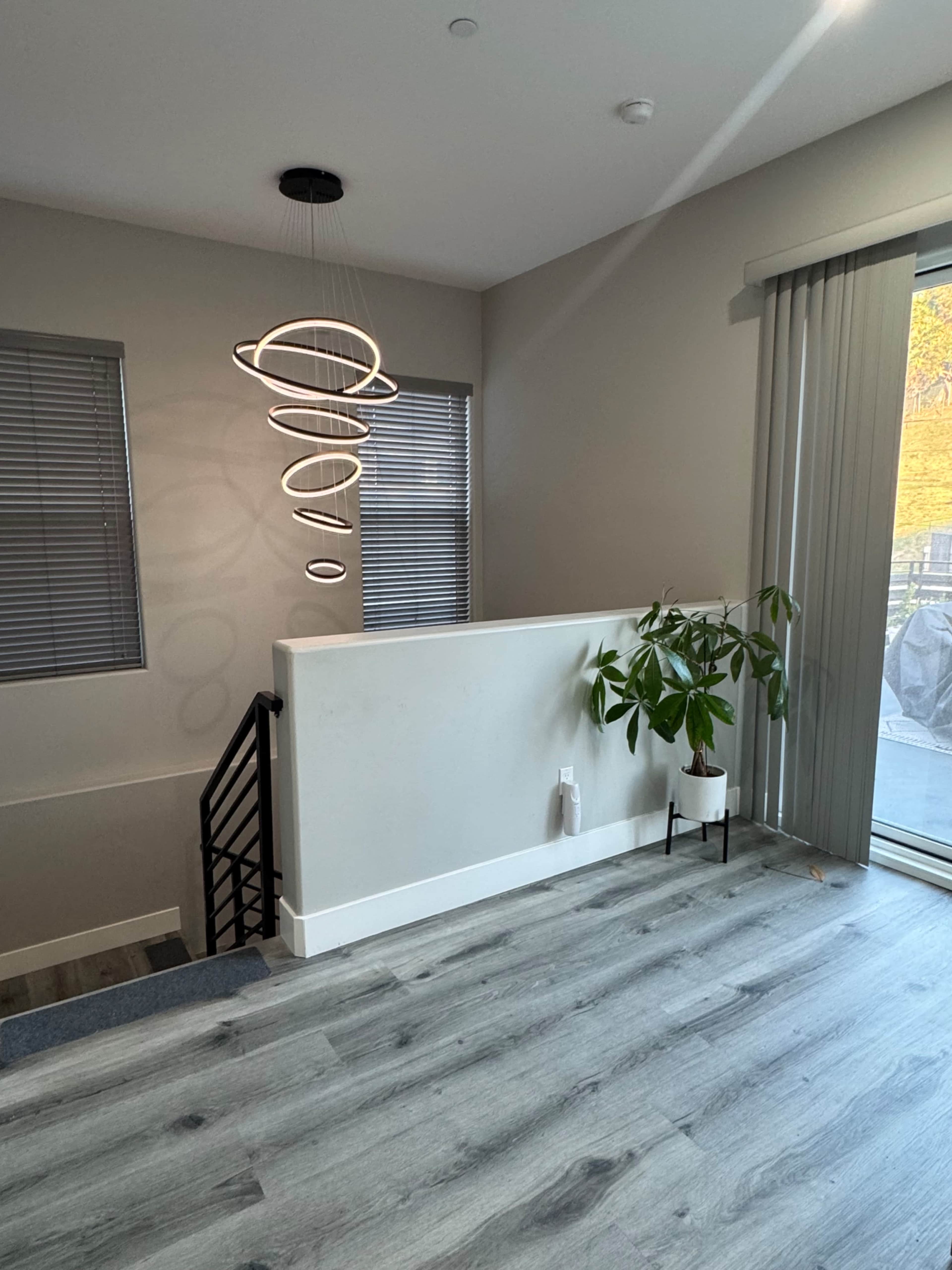The image features a well-lit, modern interior space with a spiral light fixture, a staircase, and a potted plant against a wall near large windows.