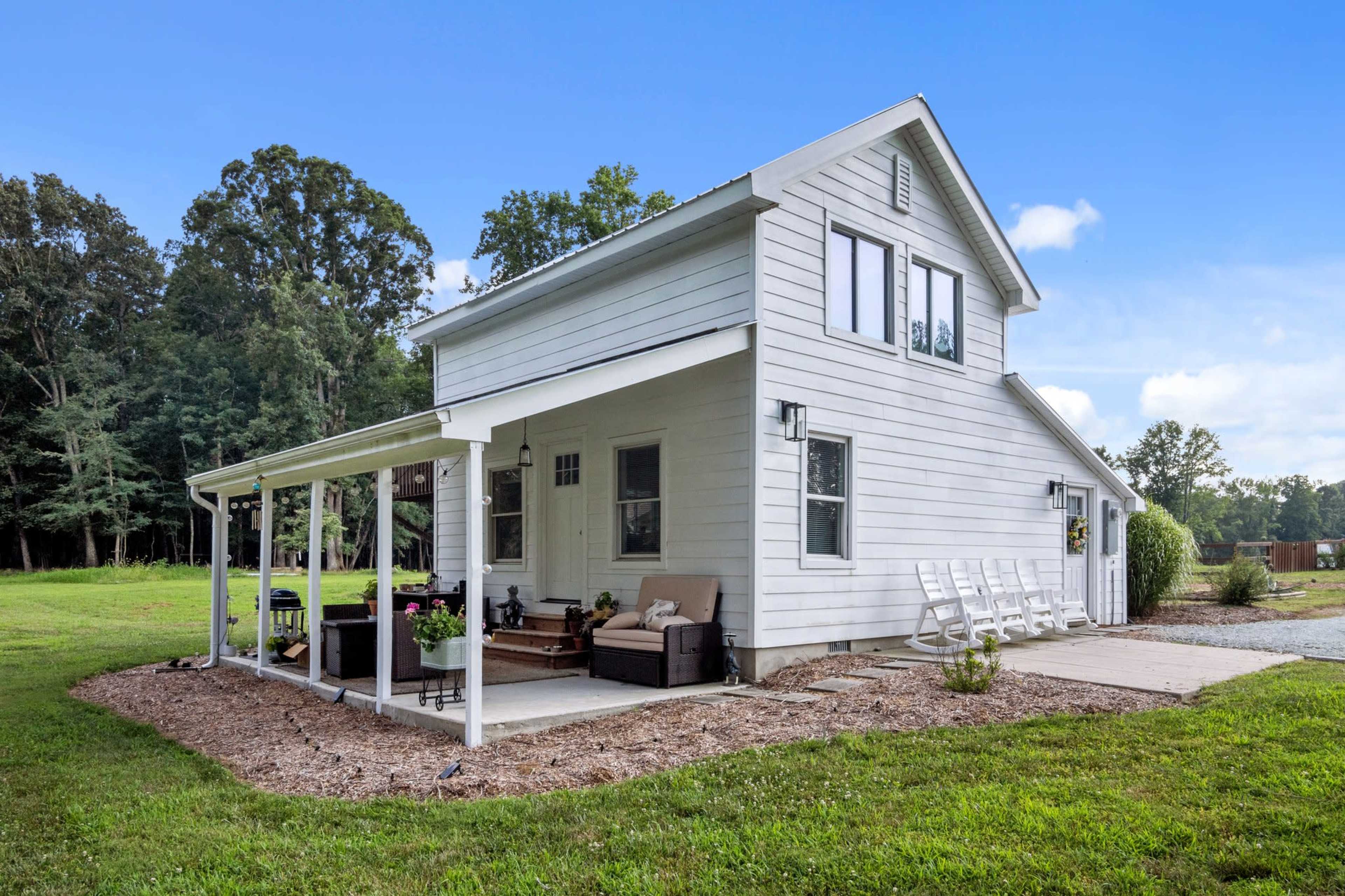 A white two-story house with a covered porch and a gravel driveway is situated on a grassy lot surrounded by trees.