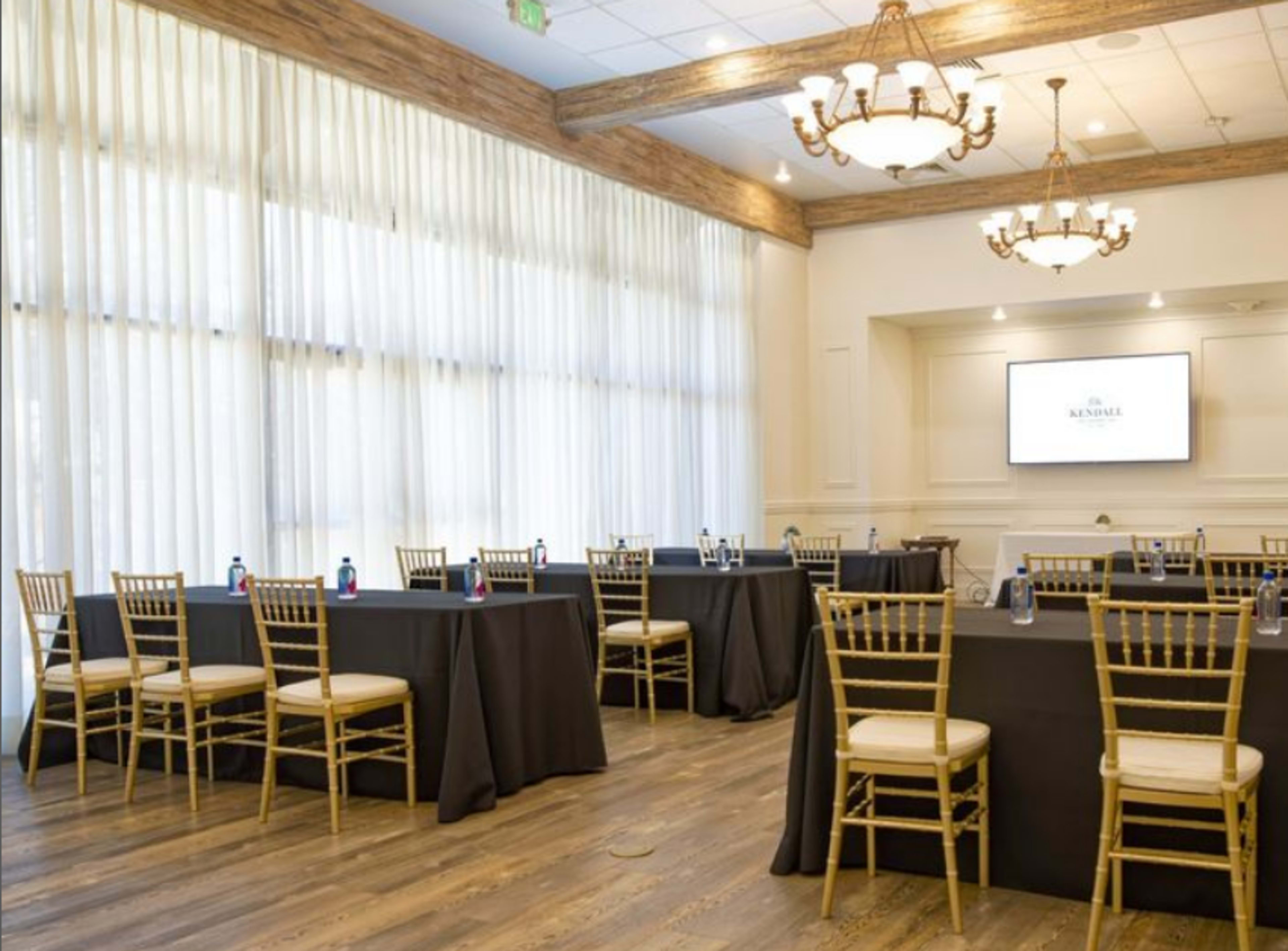 The image shows a well-lit meeting room with several round tables covered in black cloth, each surrounded by gold chairs, and a screen mounted on the wall.