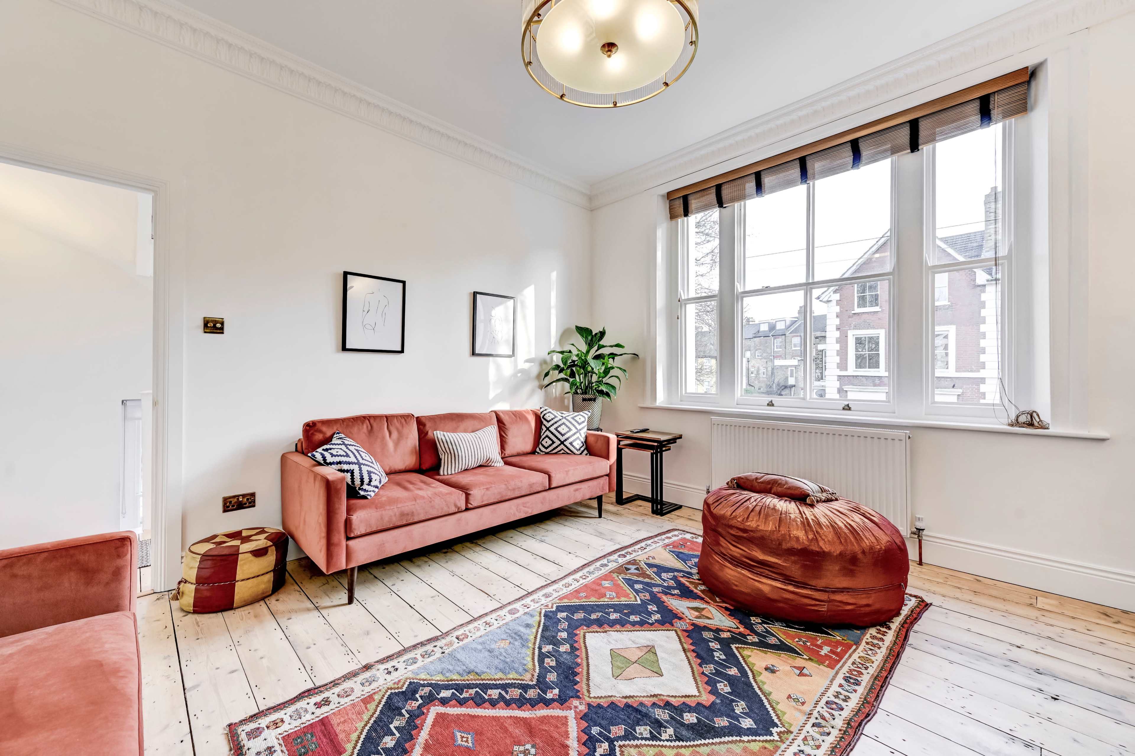 A bright living room featuring a coral sofa, a bean bag, a patterned rug, and a large window with wooden blinds.