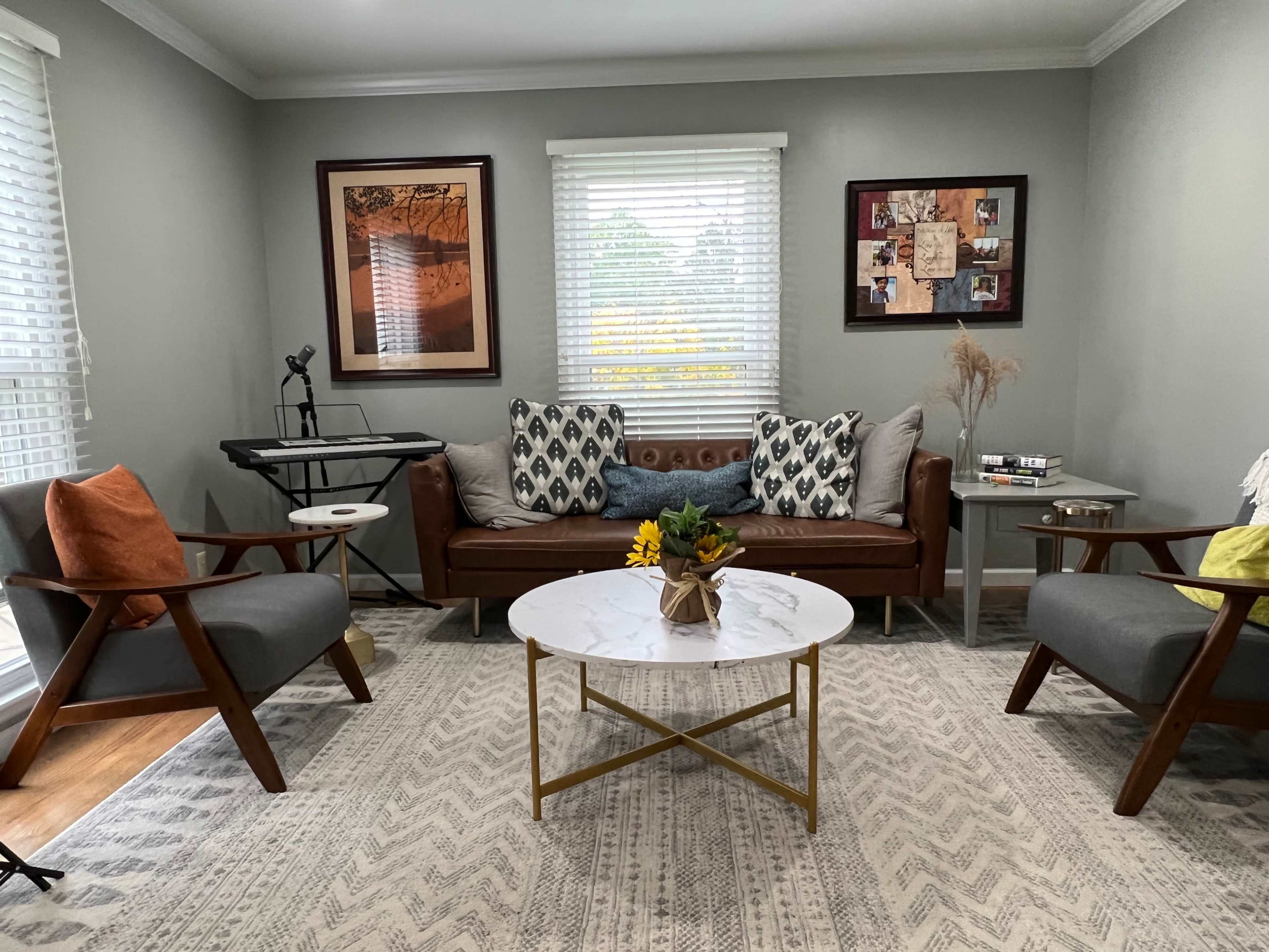 A cozy living room features a brown leather sofa with patterned throw pillows, two gray armchairs, a white marble coffee table, and a wall adorned with framed artwork.