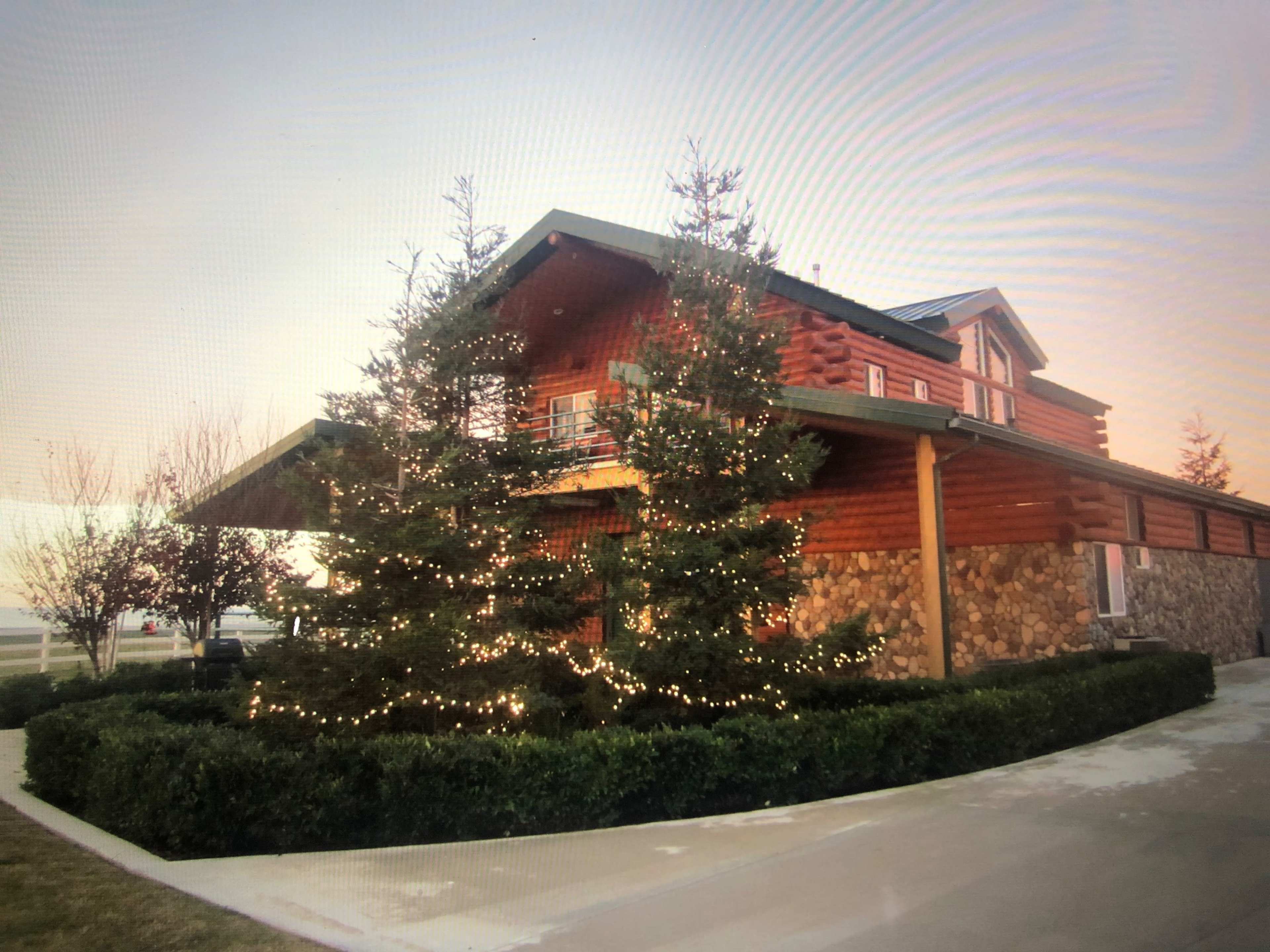 A large log cabin-style house with stone accents is surrounded by neatly trimmed bushes and two decorated evergreen trees.