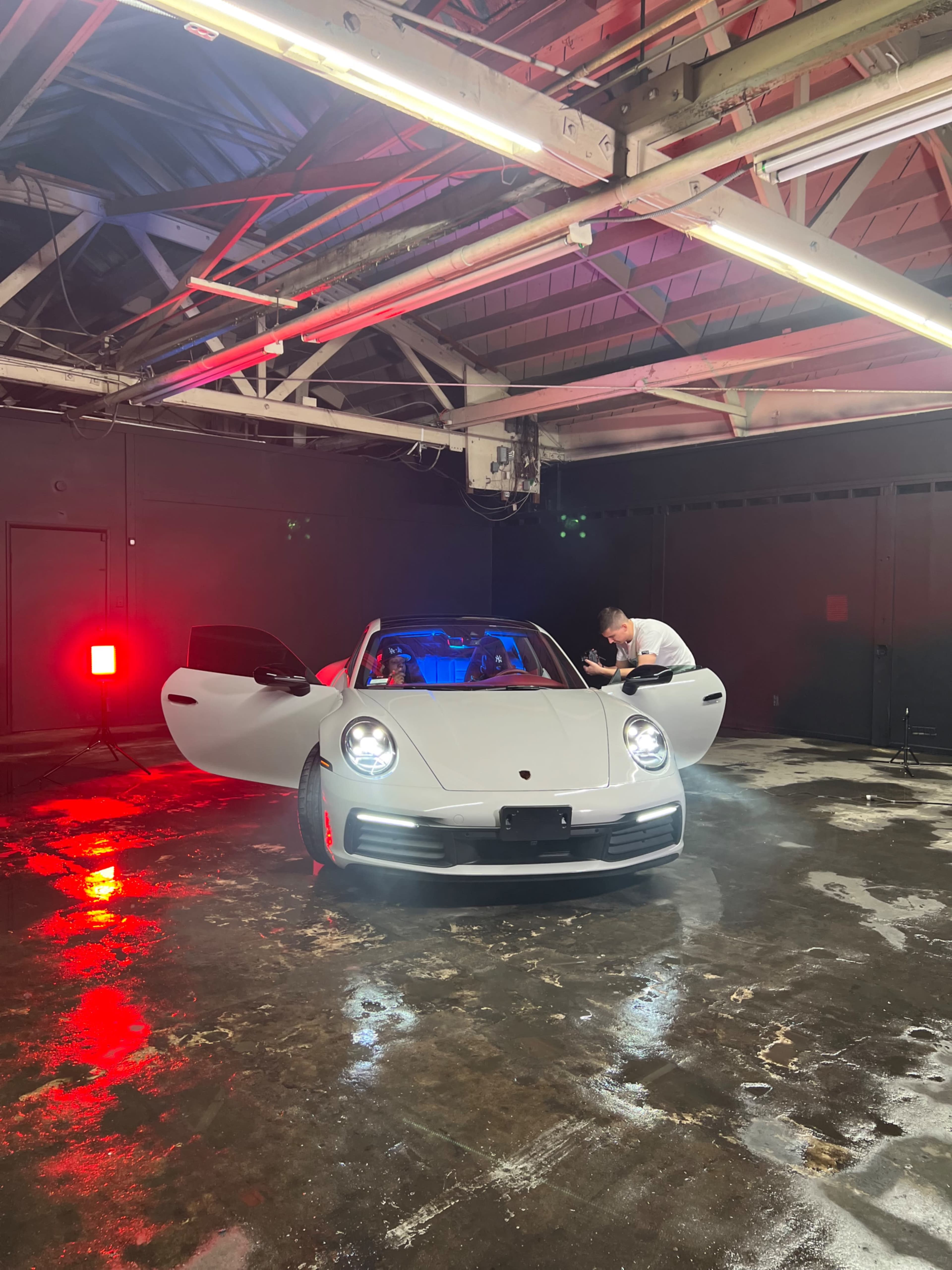 A silver Porsche 911 with its doors open is positioned in a dimly lit garage, while a person adjusts equipment nearby under colored lights.