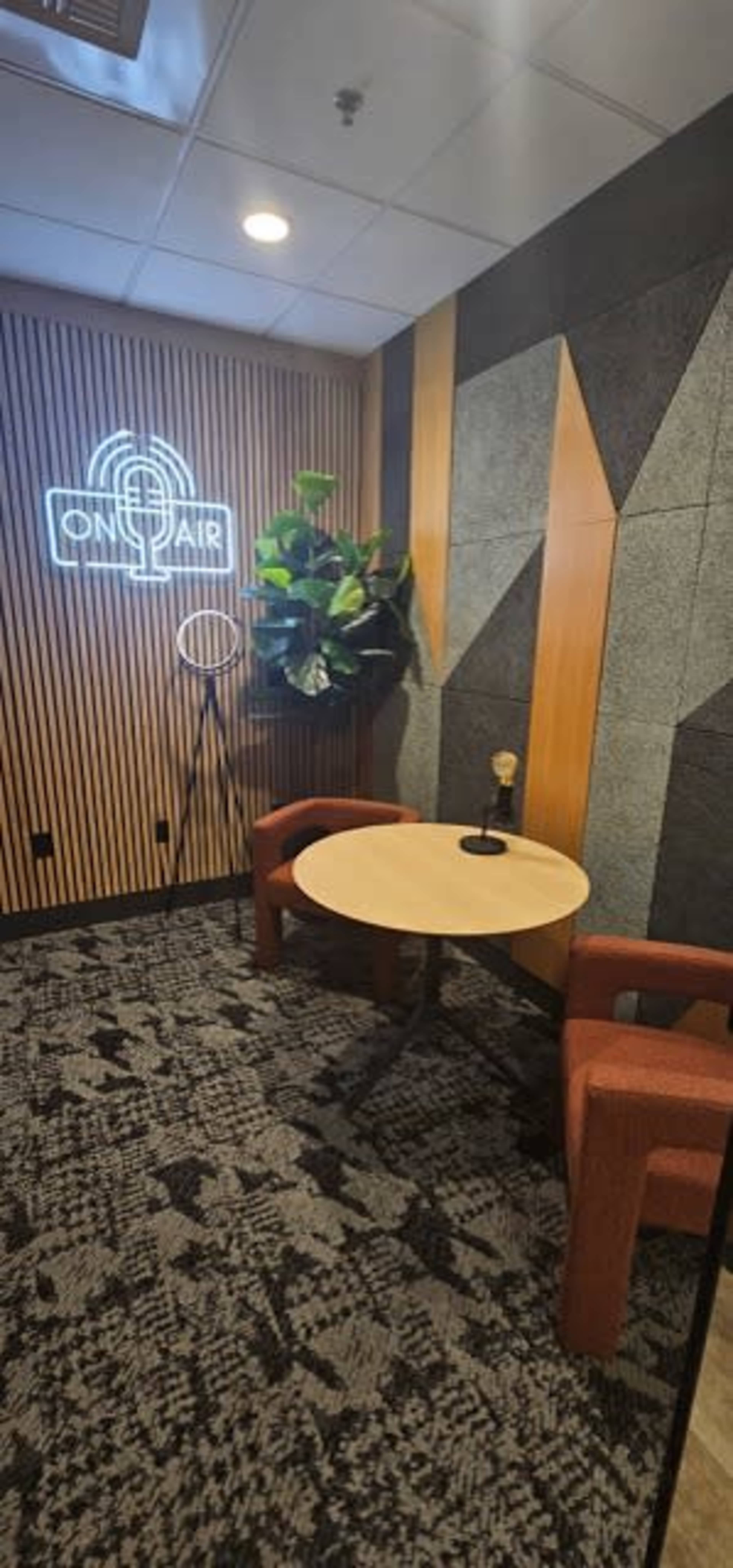 The image shows a small studio space featuring a round table with two red chairs, a decorative plant, and a wall displaying a neon "ON AIR" sign.