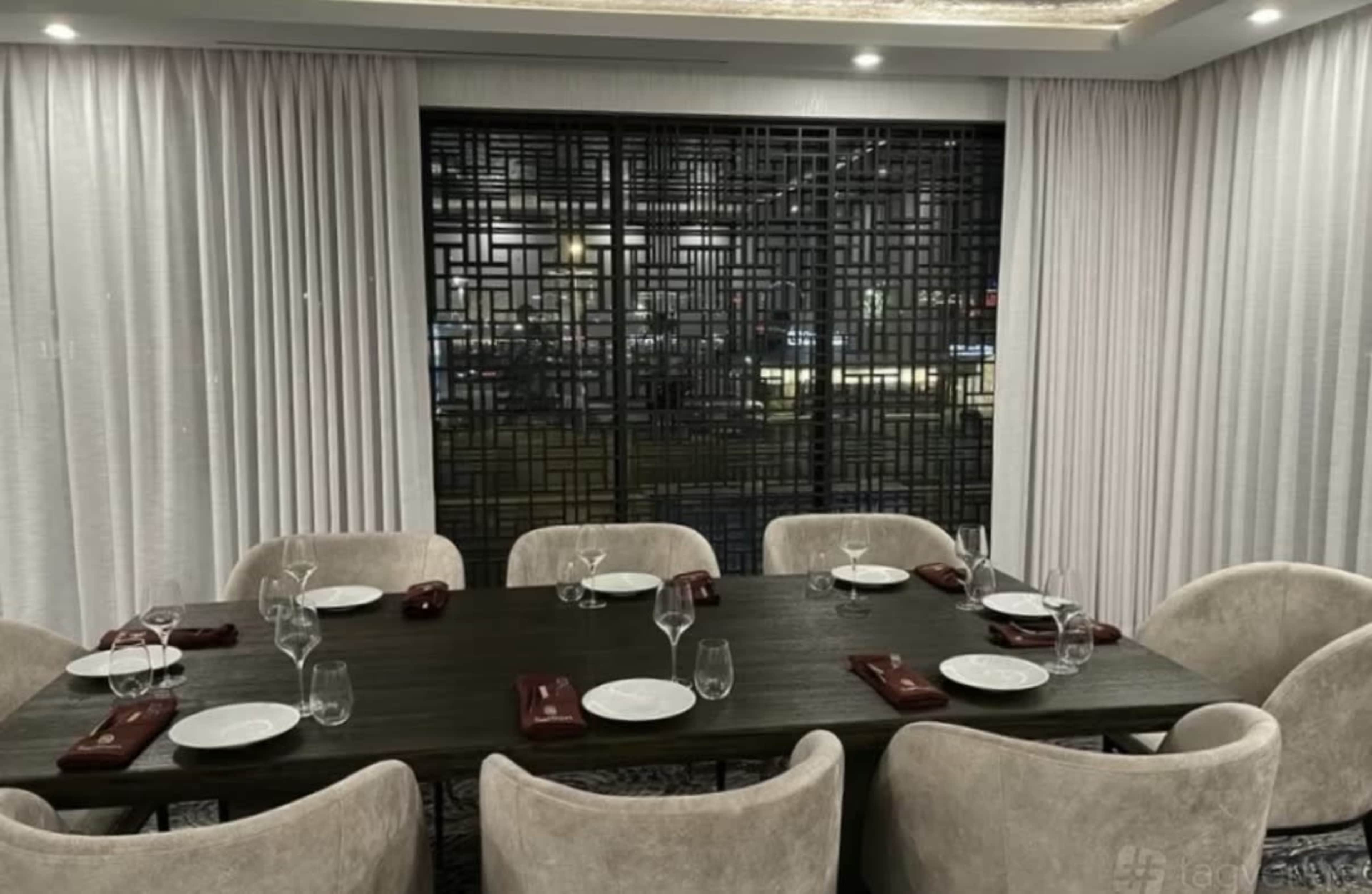 A dining table is set for eight in a well-lit restaurant with patterned glass panels and curtains in the background.