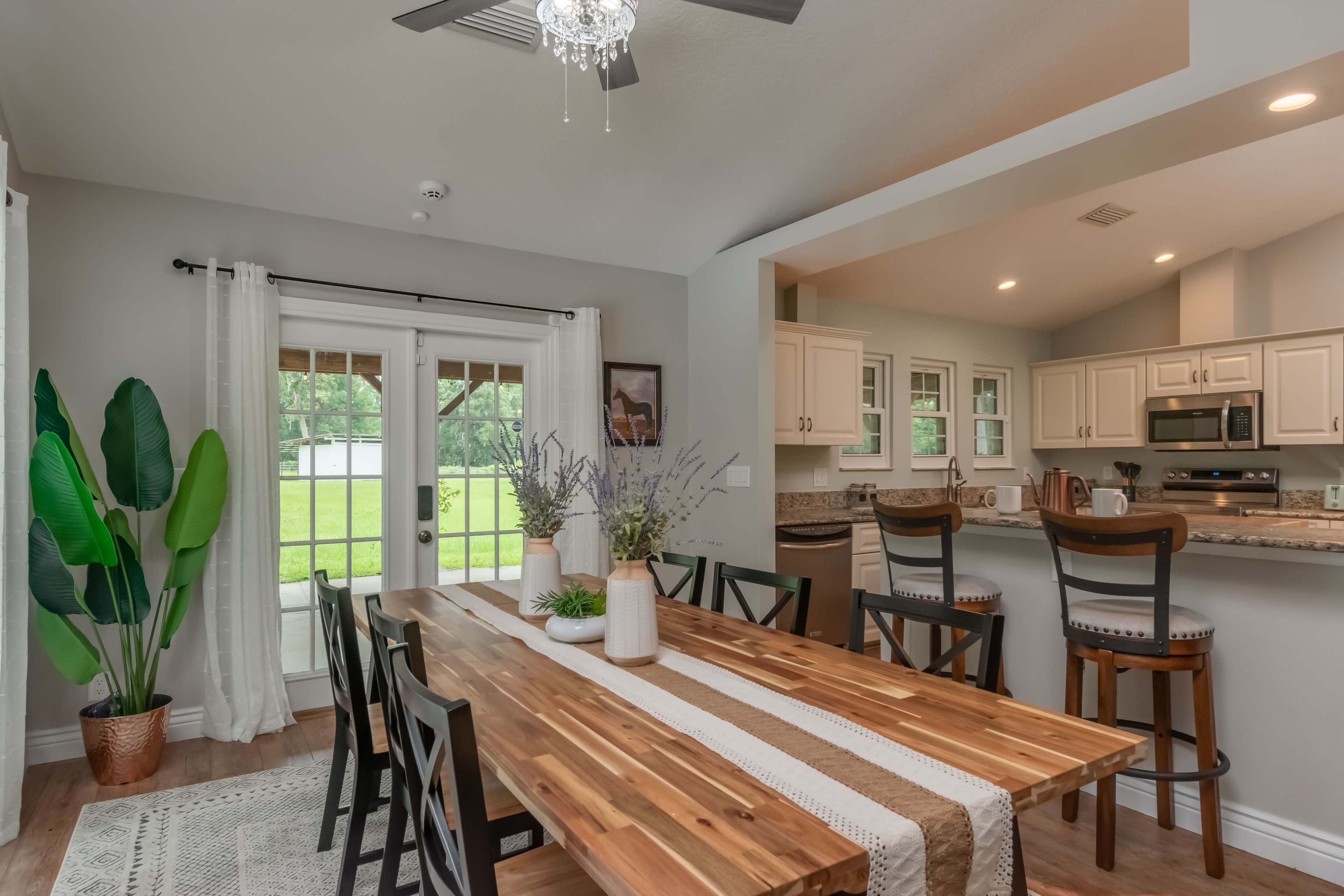 A dining area features a long wooden table surrounded by black chairs, with a view of greenery through French doors and a kitchen visible in the background.