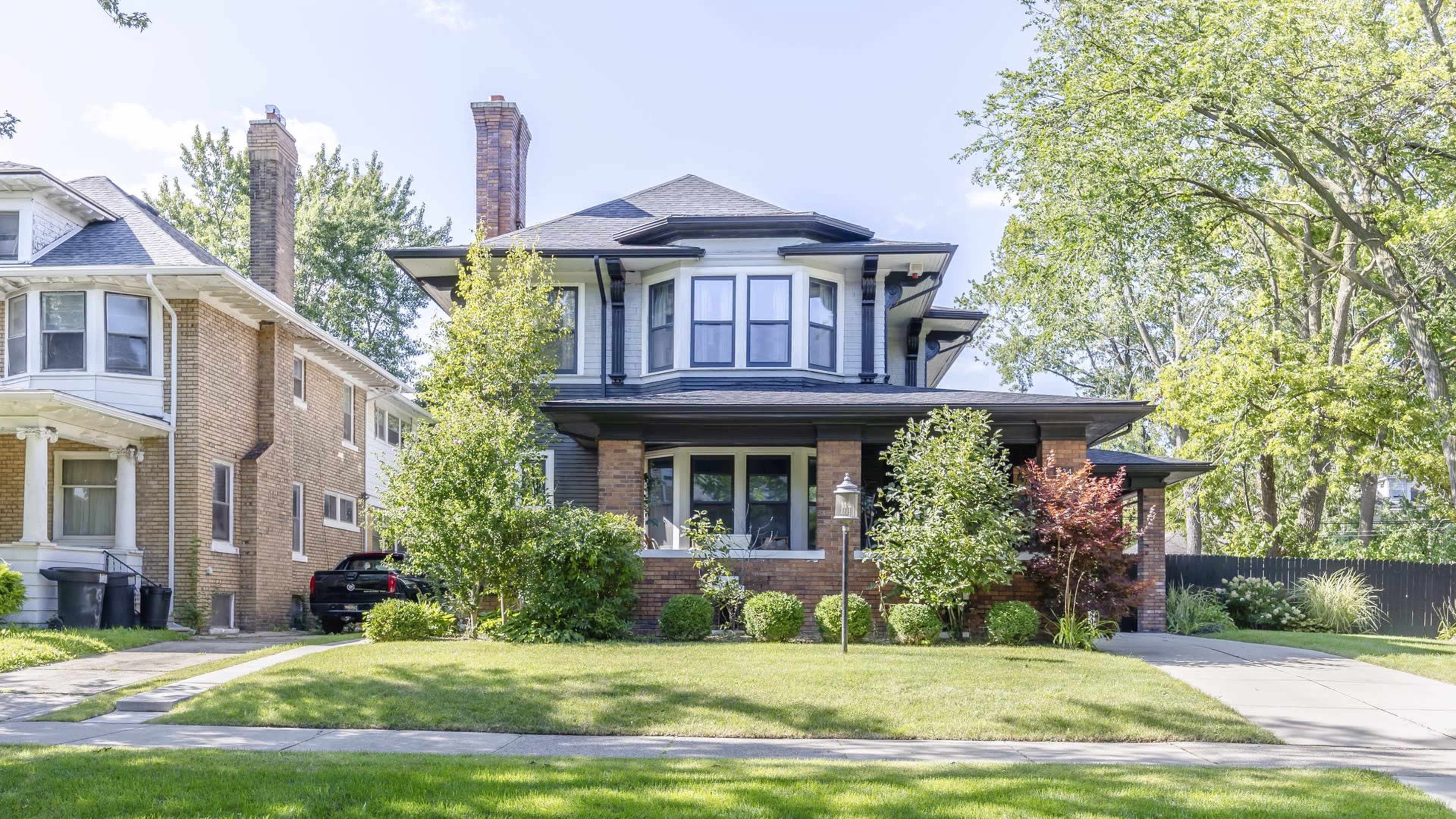 A two-story brick house with a wide porch and large bay windows stands surrounded by well-maintained shrubs and a lawn.
